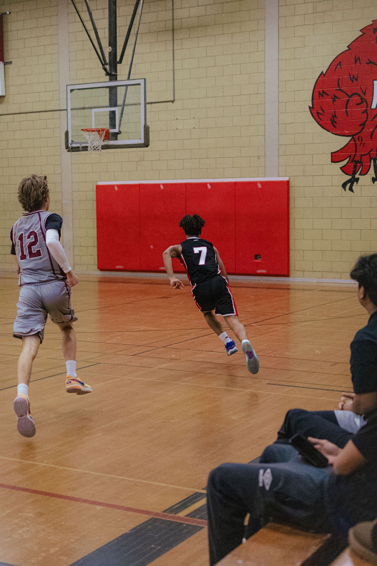 Players drive toward the basketball hoop during an indoor game with a red wall backdrop.