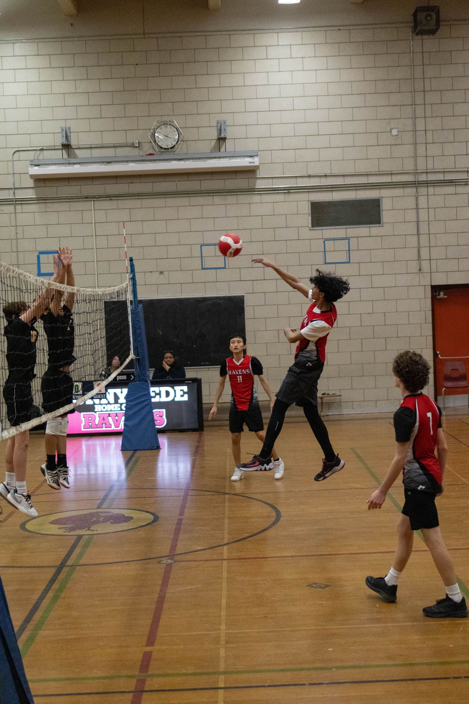 Indoor volleyball match with player in red uniform spiking ball over the net.