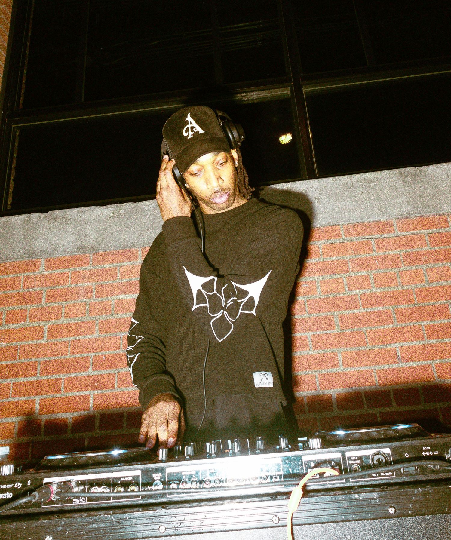 A DJ performs at night wearing a black hoodie and baseball cap while using turntables.
