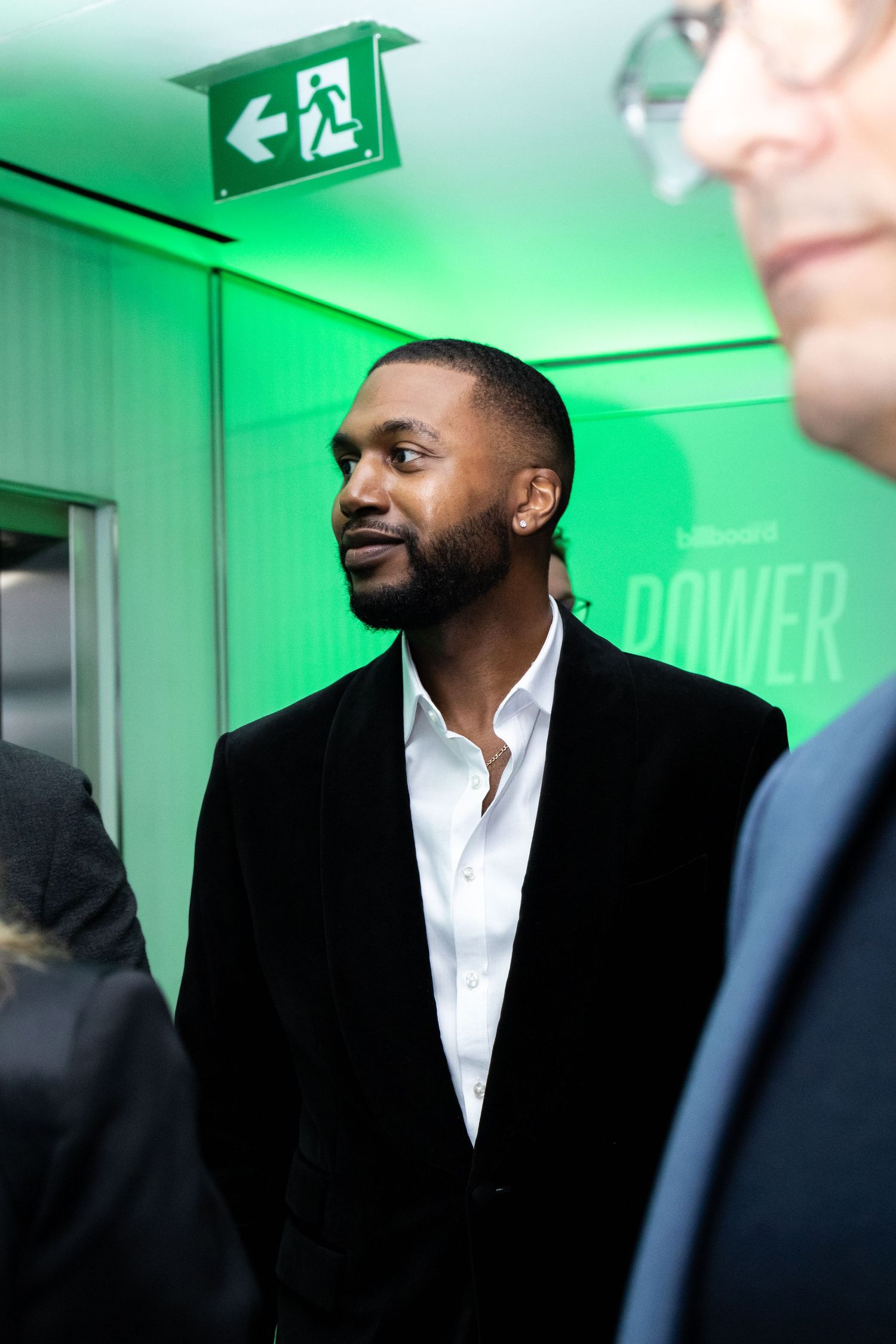 Man in black suit and white shirt stands in hallway with green exit sign lighting.