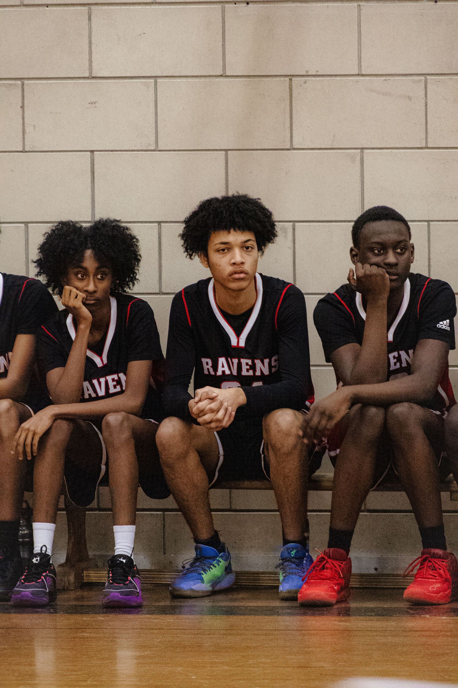 Three athletes in Ravens basketball uniforms sit on a bench against a concrete block wall.
