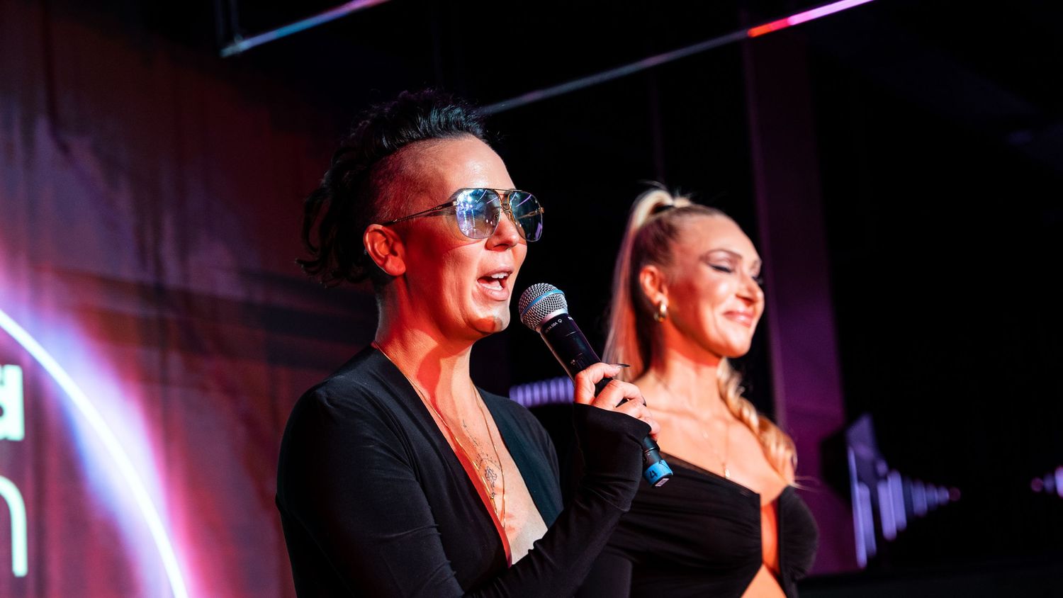Two performers singing together enthusiastically on stage under colorful lighting.