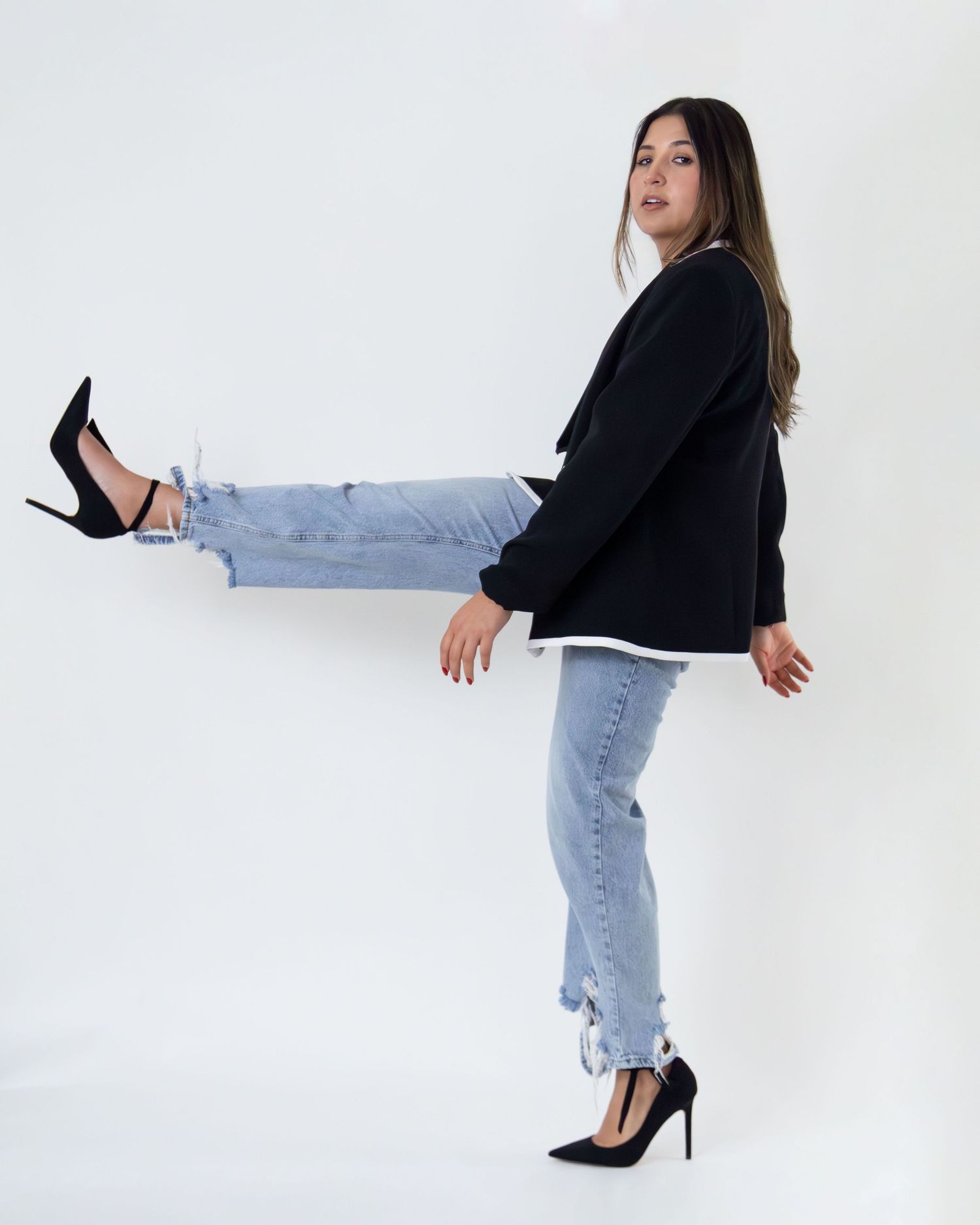 Woman in black blazer, white top, light wash jeans, and black heels posing with one leg raised on white background.