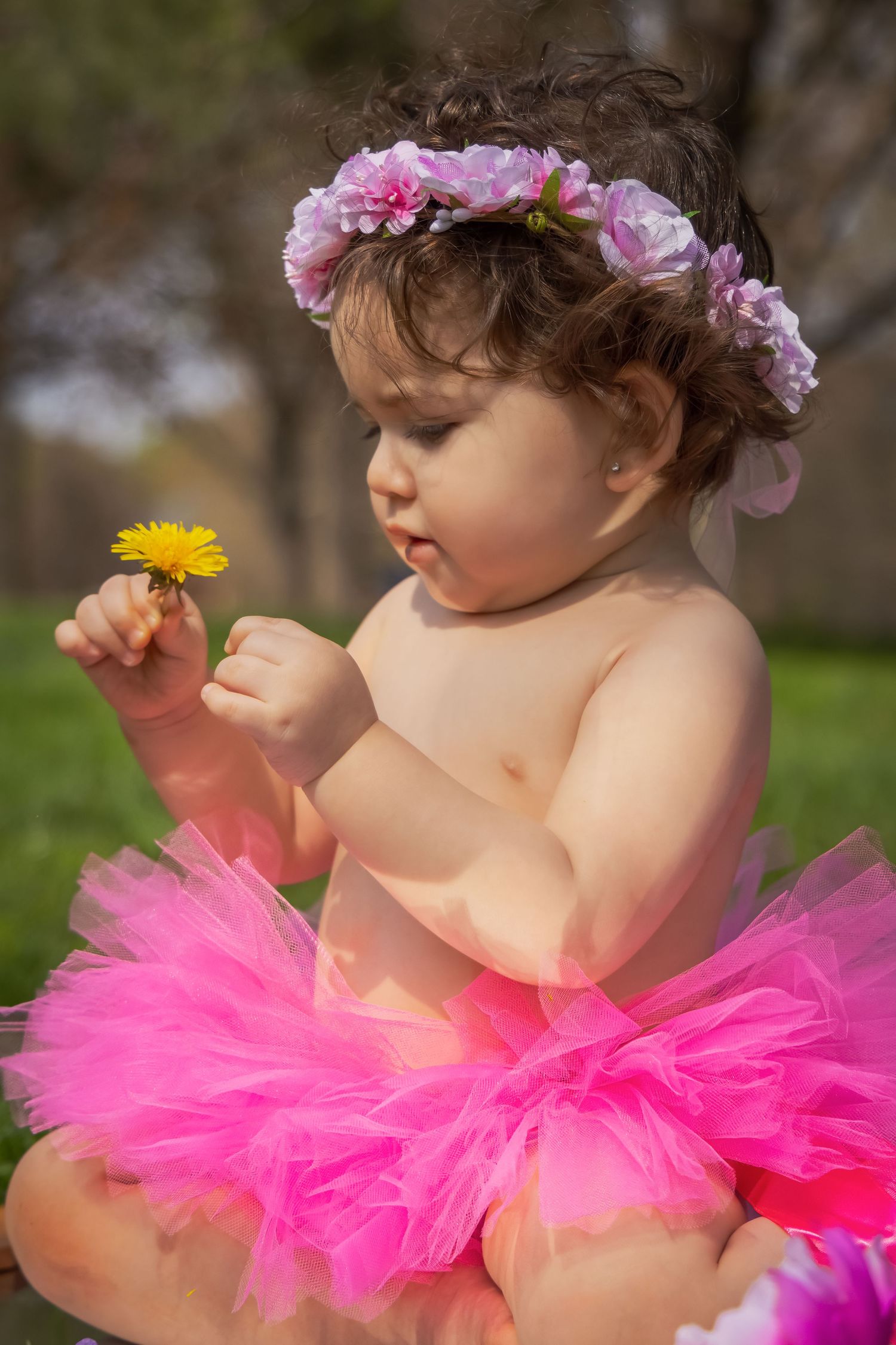 Baby girl in pink tutu and floral crown holds a yellow dandelion outdoors in a sunny garden setting.