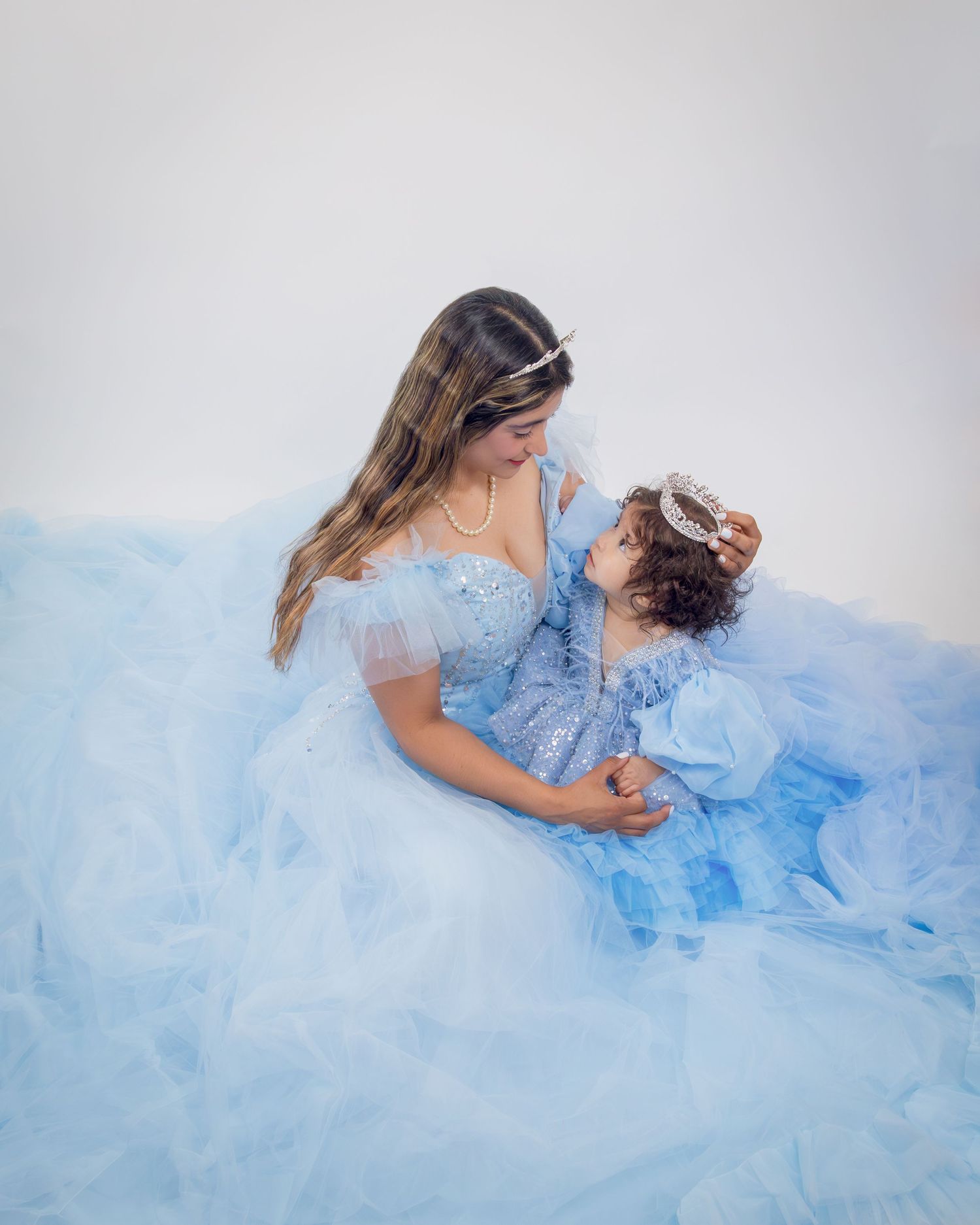 A woman and toddler in matching light blue tulle gowns share a tender moment in a dreamy studio portrait.