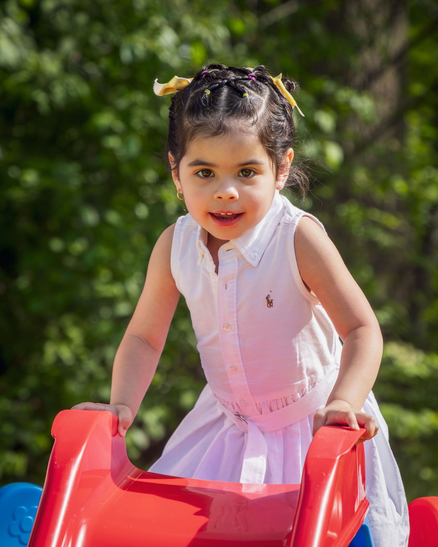 Child wearing a pink dress plays on a bright red playground toy on a sunny day, surrounded by greenery.