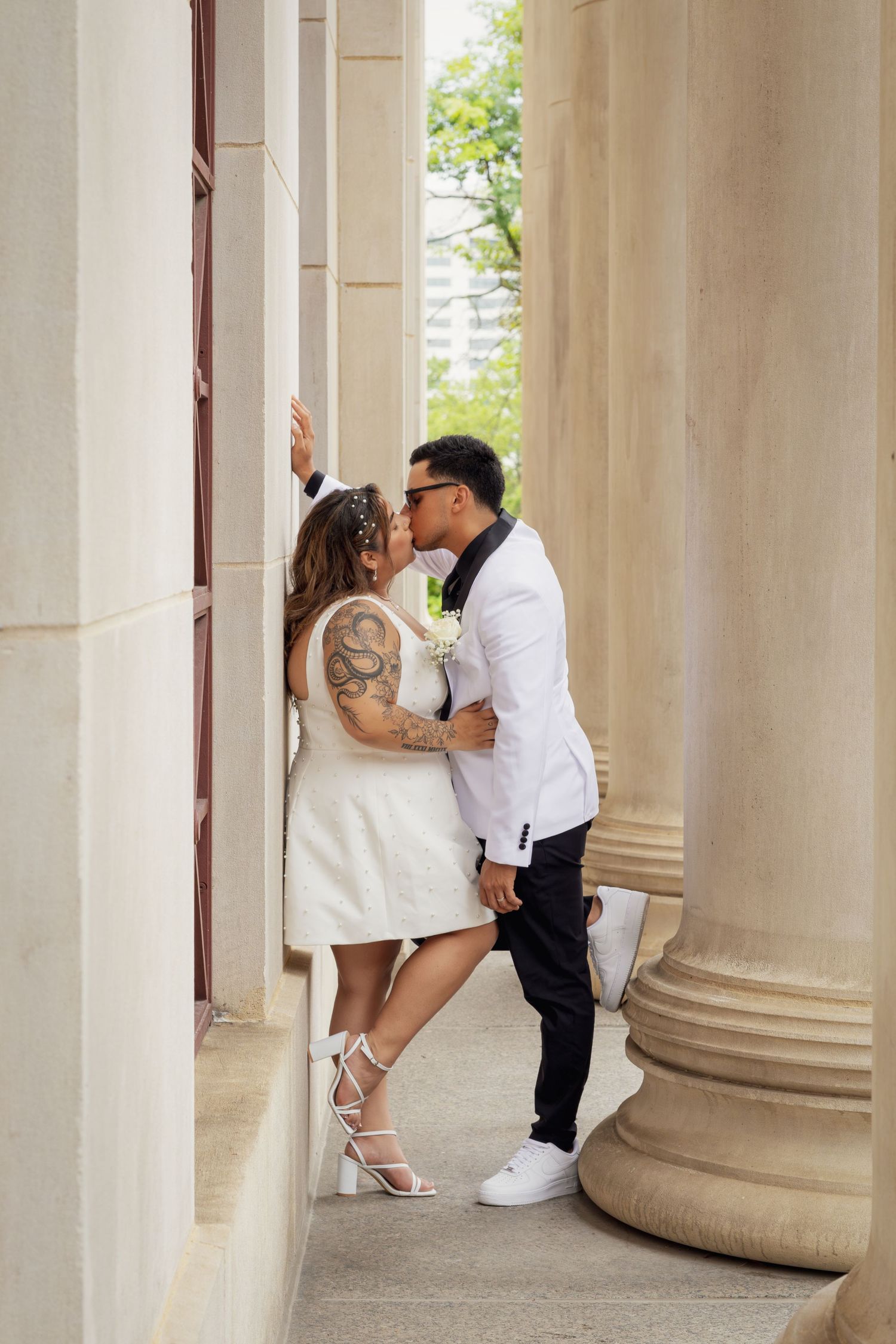 Couple sharing a romantic kiss between elegant stone columns, bride in white mini dress with tattoos, groom in white blazer.