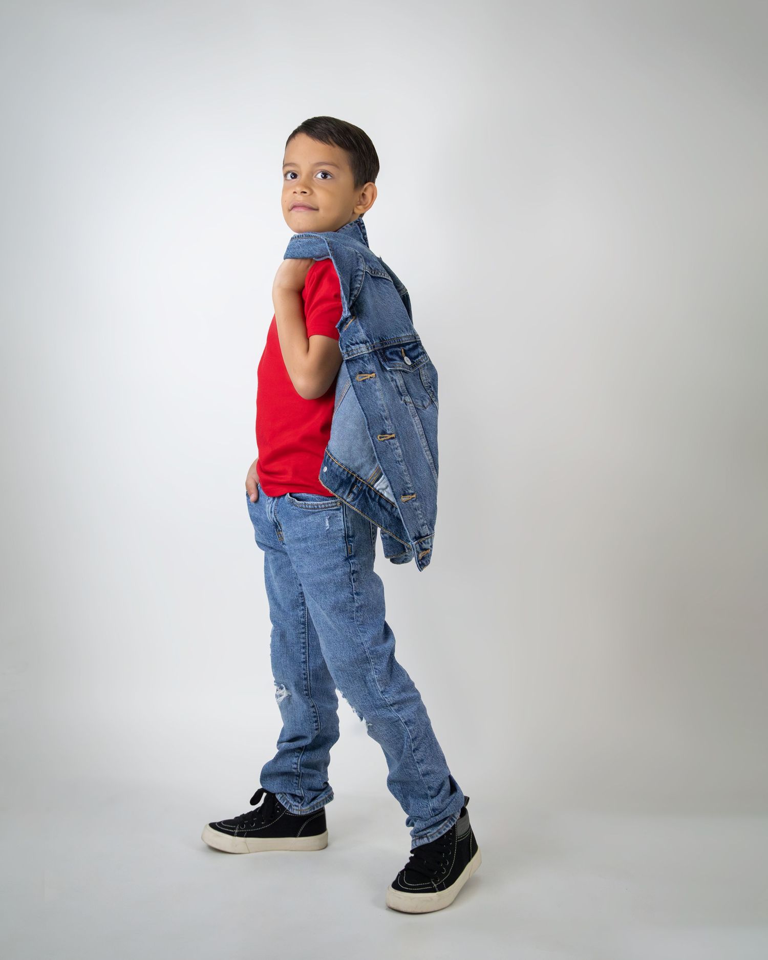 A young boy poses in a red shirt, ripped jeans, and black sneakers, holding a denim jacket over his shoulder.