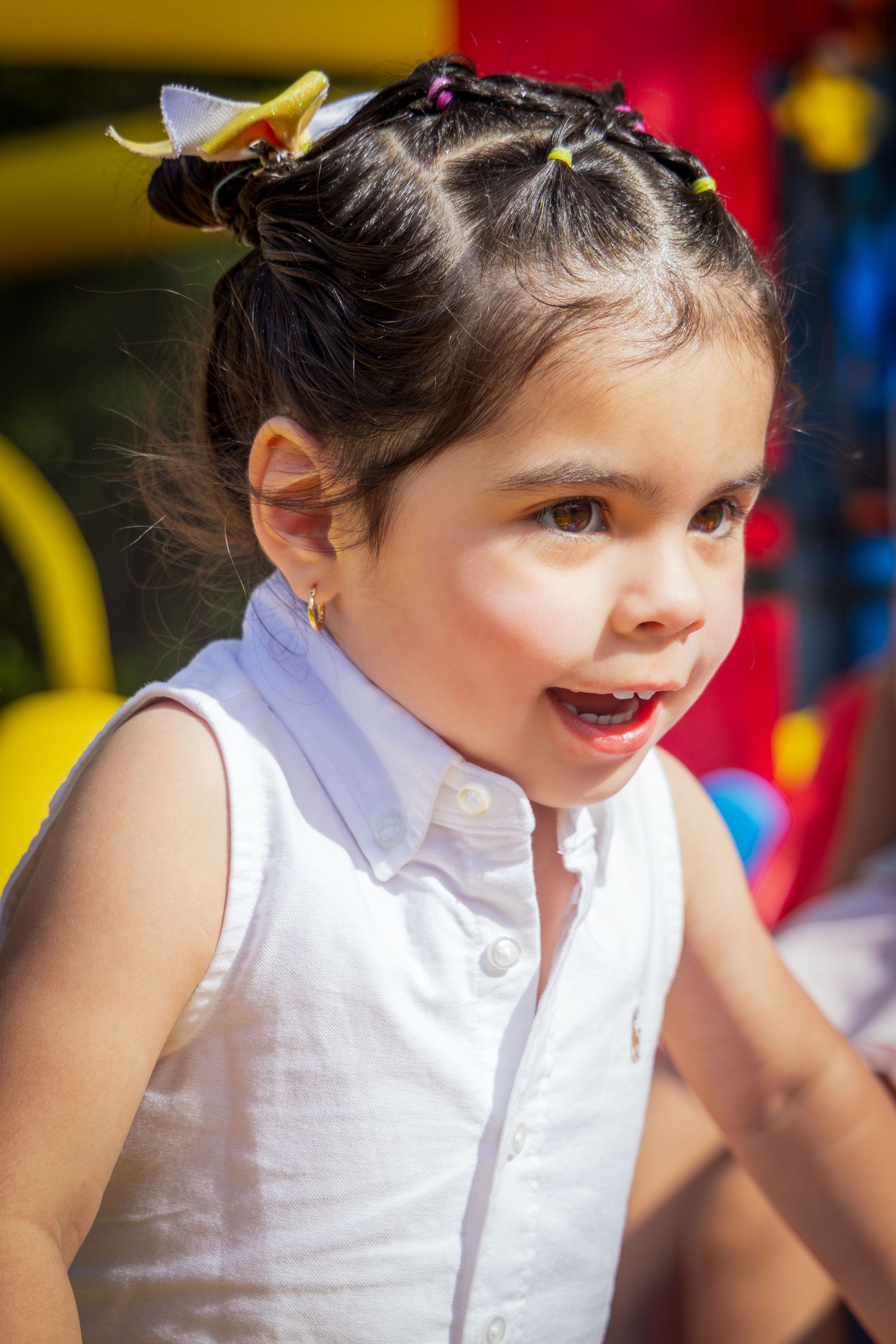 Young girl with braided hair and white sleeveless shirt smiling outdoors at a colorful playground.