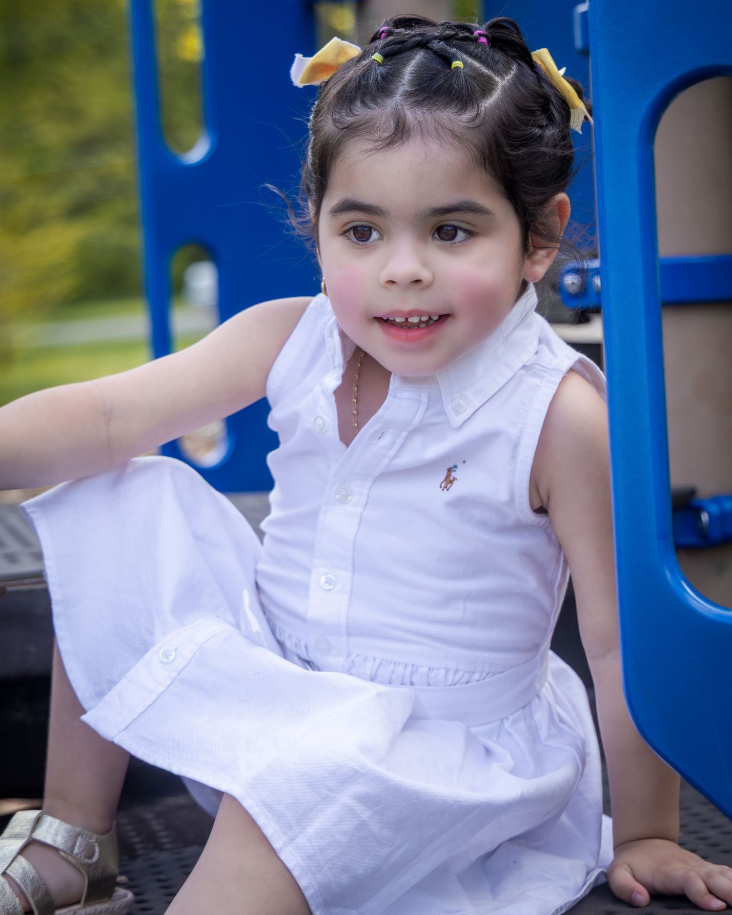 Child in a crisp white sleeveless dress sitting on blue playground equipment on a sunny day.