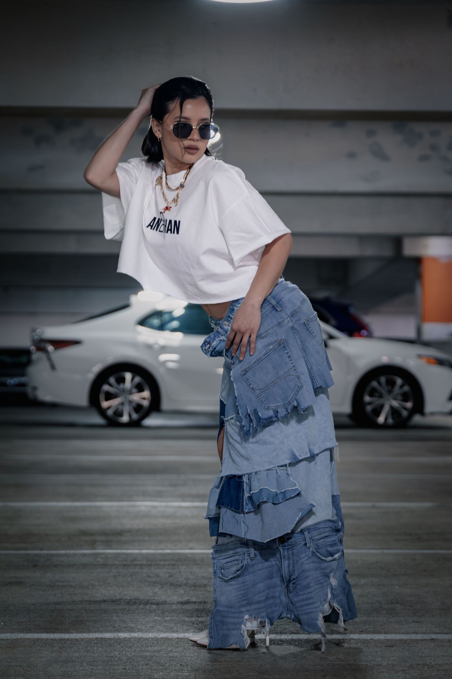 Woman in white Jordan crop top and layered denim skirt posing in parking garage with sunglasses.