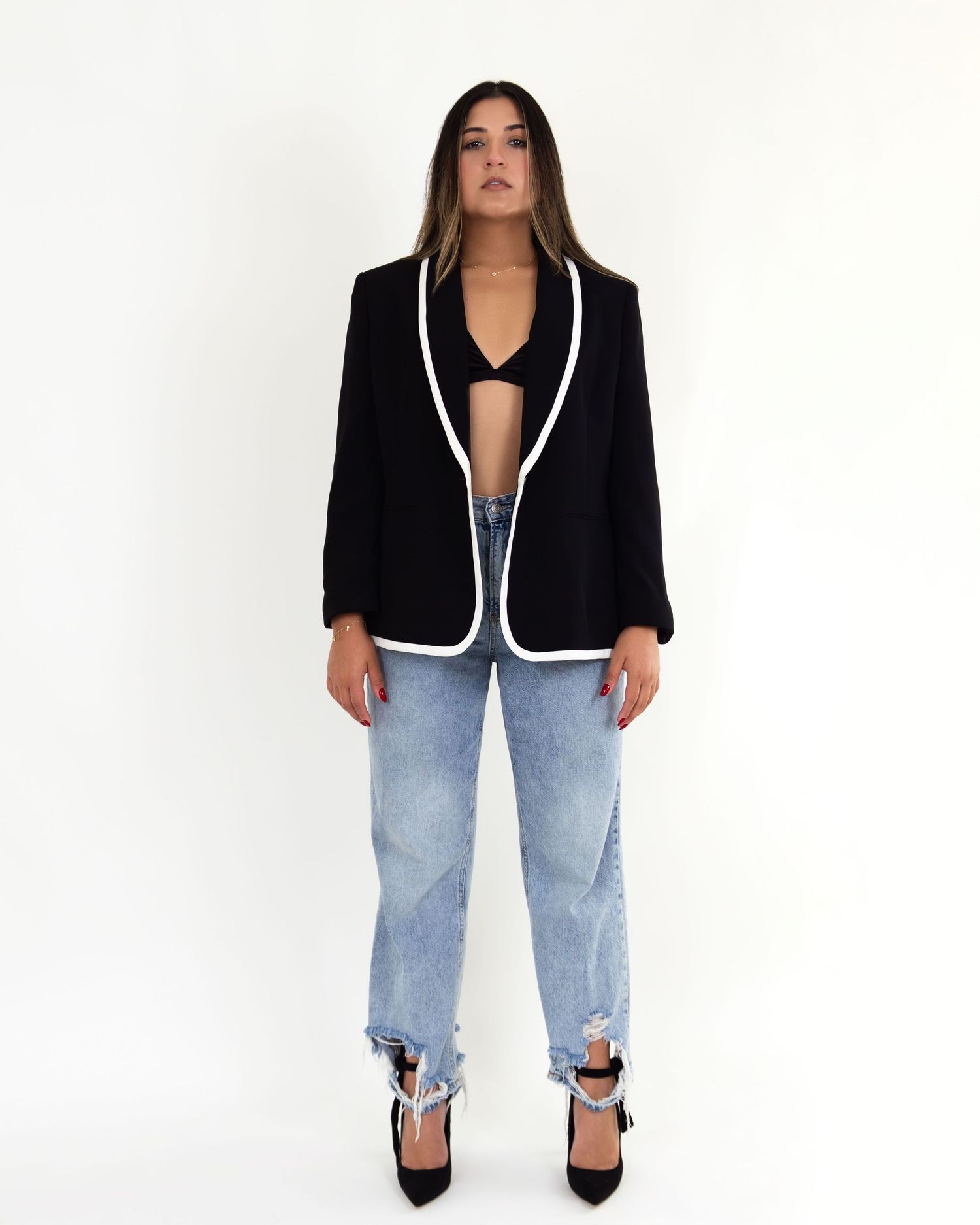 Woman wearing black blazer with white trim, black bralette, light wash ripped jeans, and black heels.