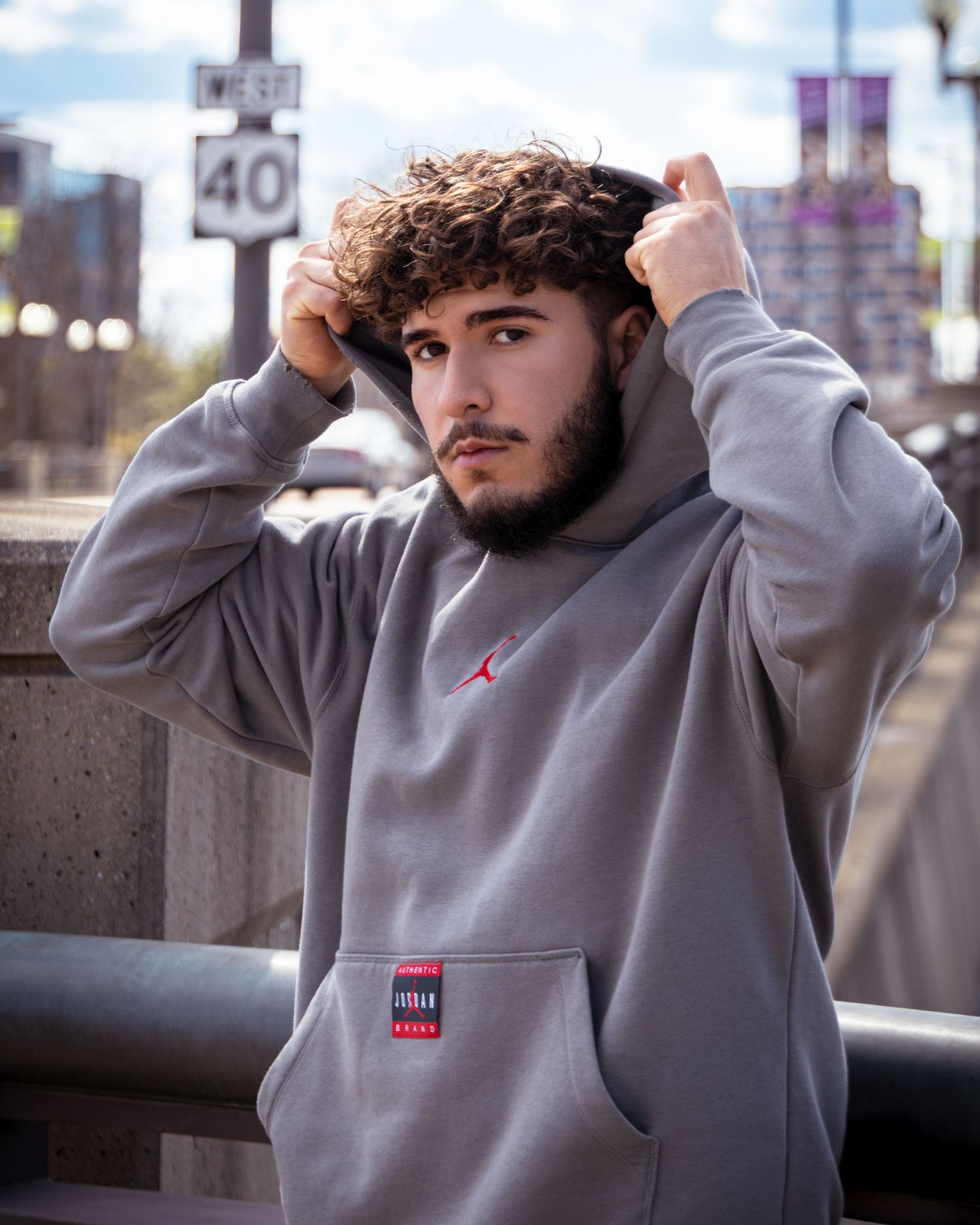 Young man with curly hair wearing a gray Jordan Brand hoodie poses on a city bridge near a speed limit sign.