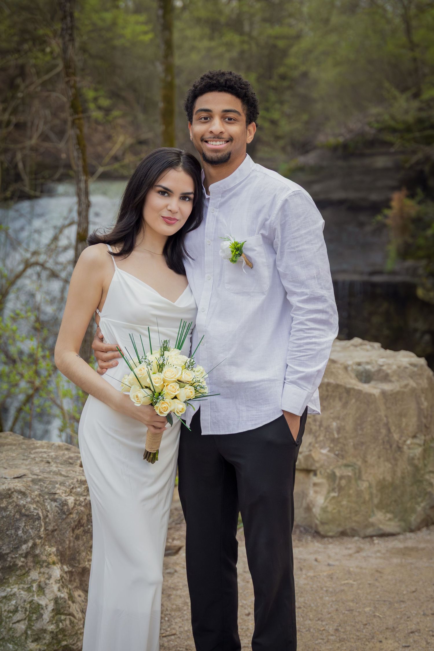 A smiling interracial couple poses outdoors in wedding attire, bride holding yellow roses bouquet near a forest waterfall.
