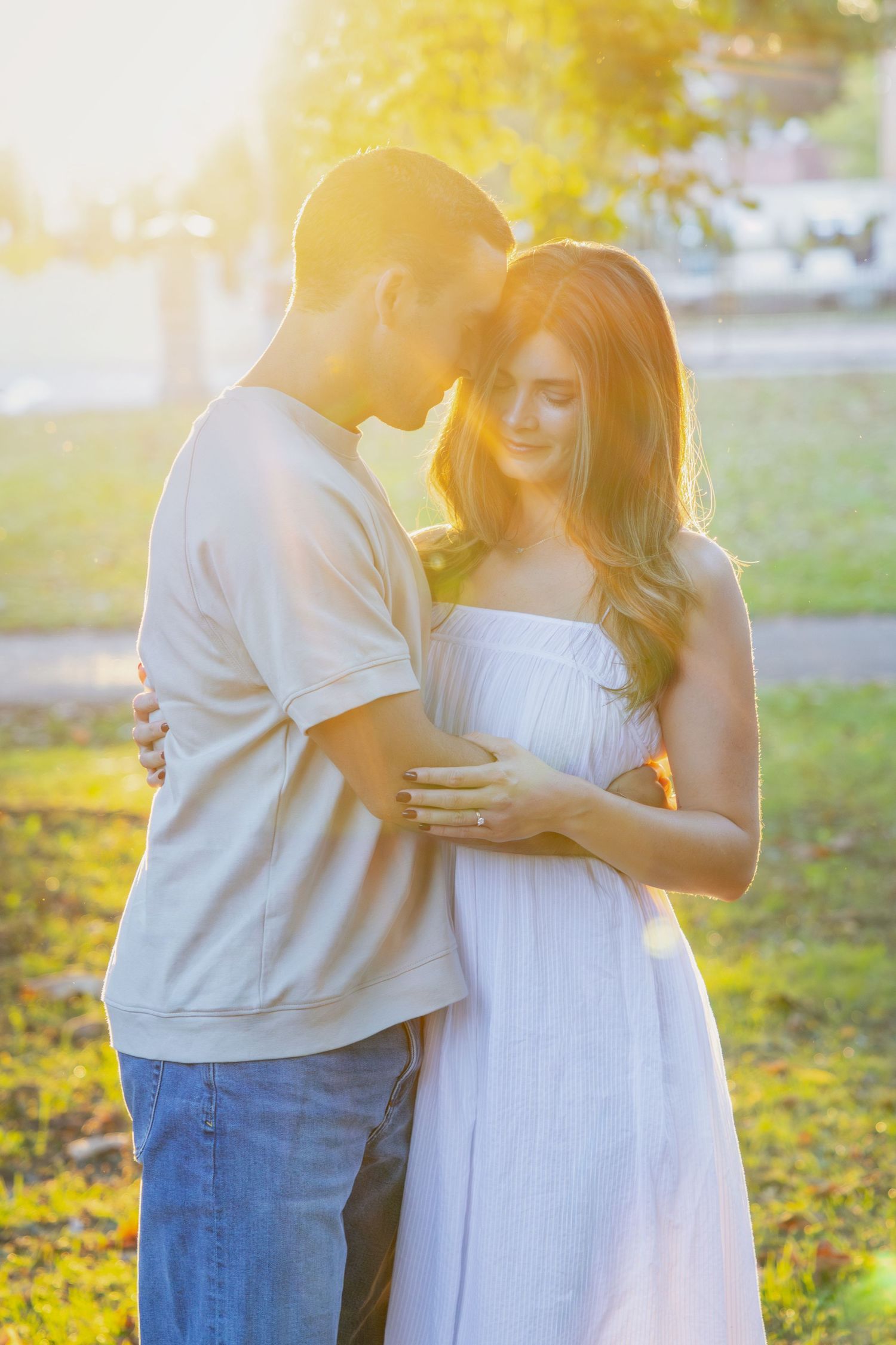 A couple embraces in a sunny park with golden sunlight filtering through trees in a romantic outdoor setting.
