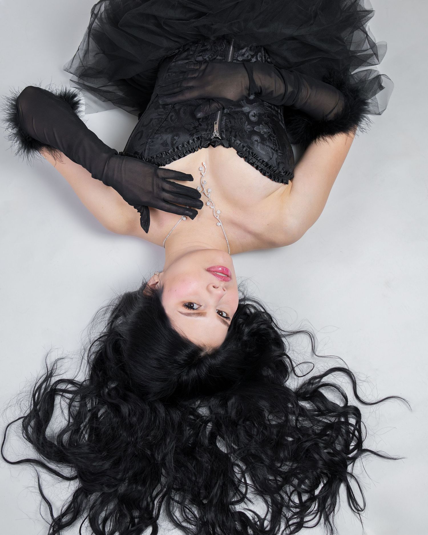 A woman in black lace corset and long gloves lies on a white floor with flowing dark hair spread around her.