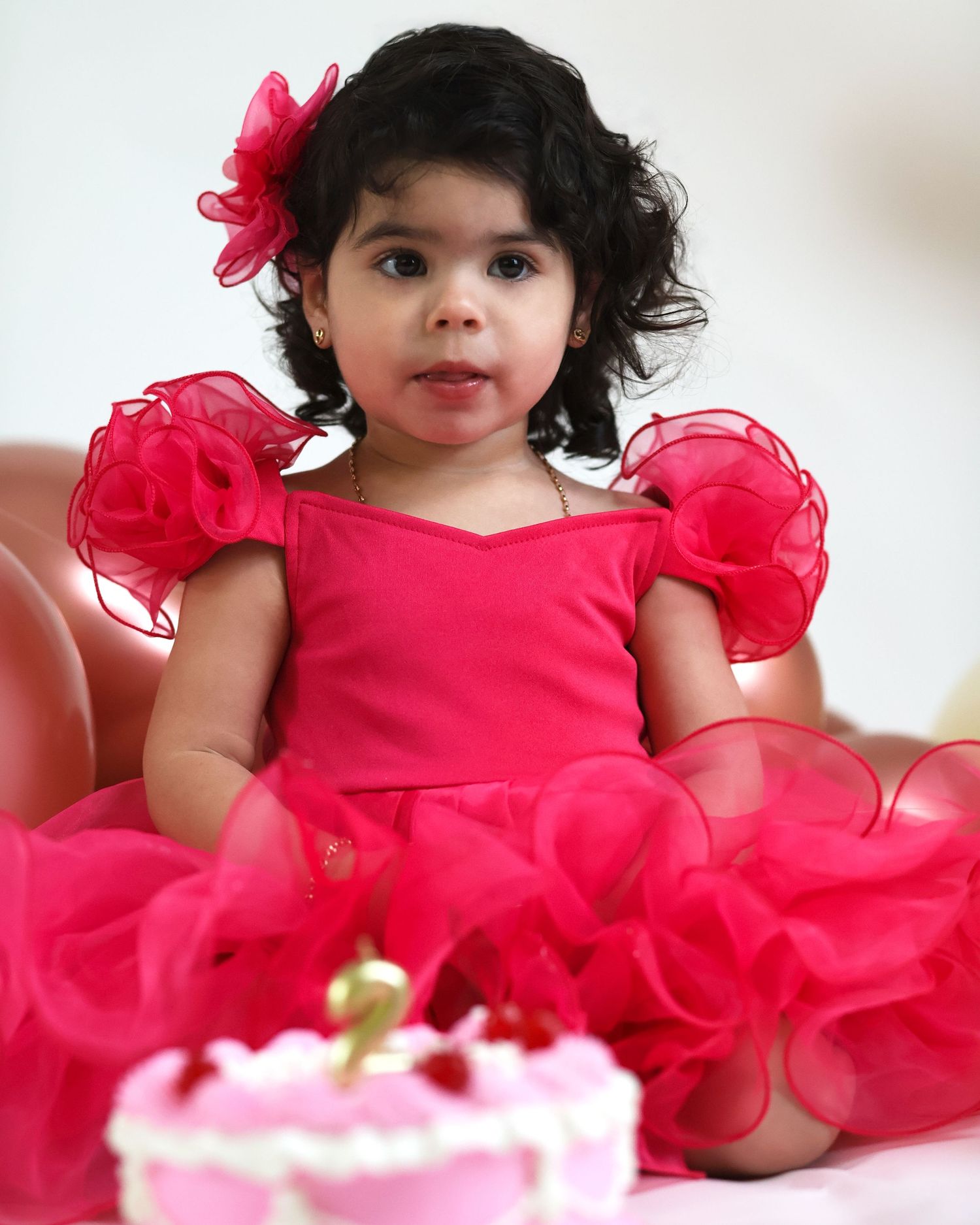 Toddler wearing red ruffled party dress smiles while seated with pink birthday cake.
