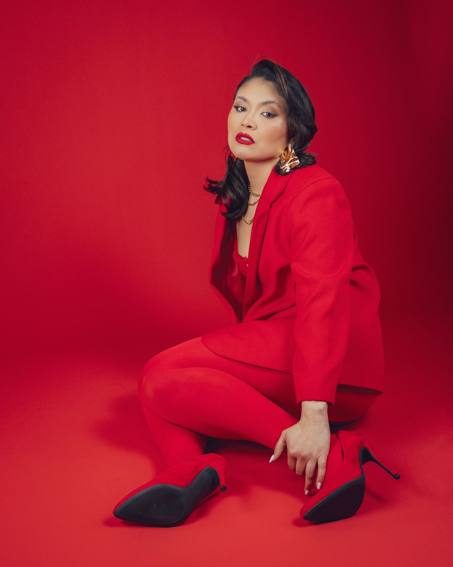 Woman in red suit and heels poses on red background with gold earrings and red lips.