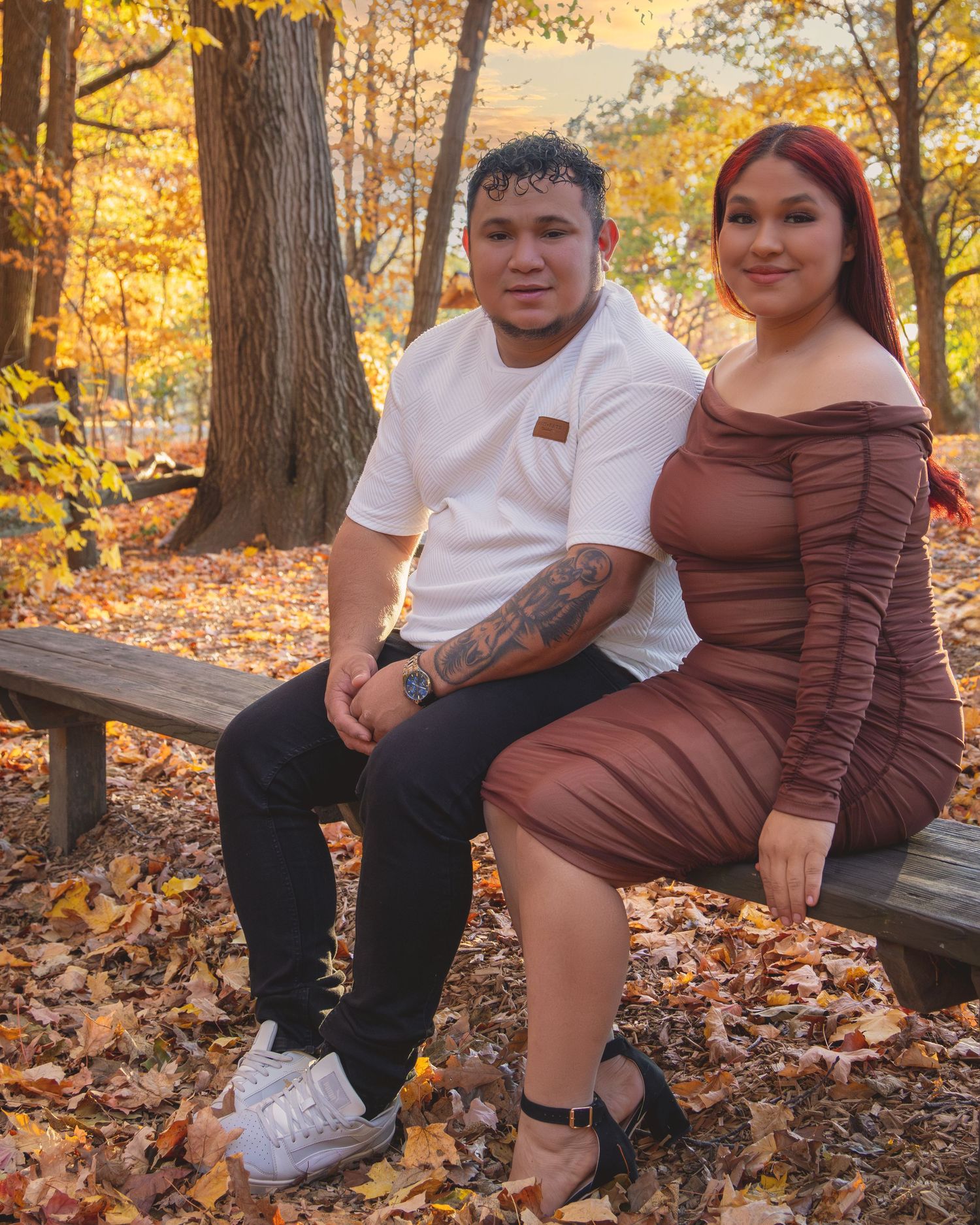 Couple sitting on a bench surrounded by autumn leaves in a park, man in white tee and woman in brown dress.