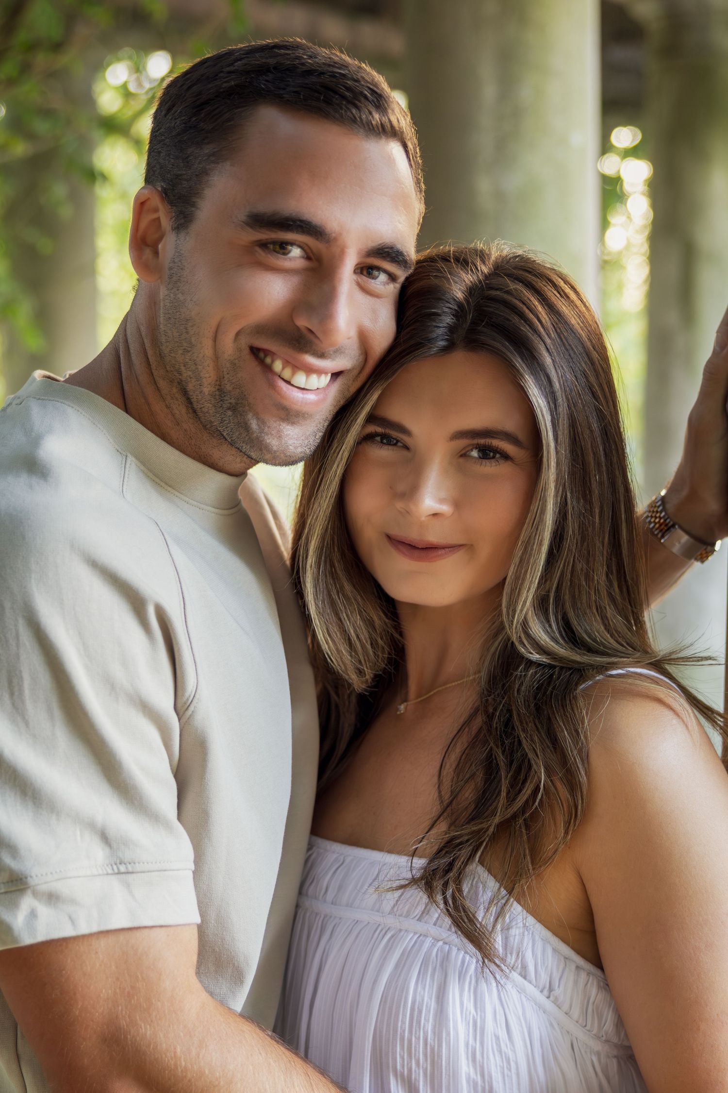 A smiling couple poses closely together outdoors, the man in a beige shirt and woman in a white dress, surrounded by lush greenery.