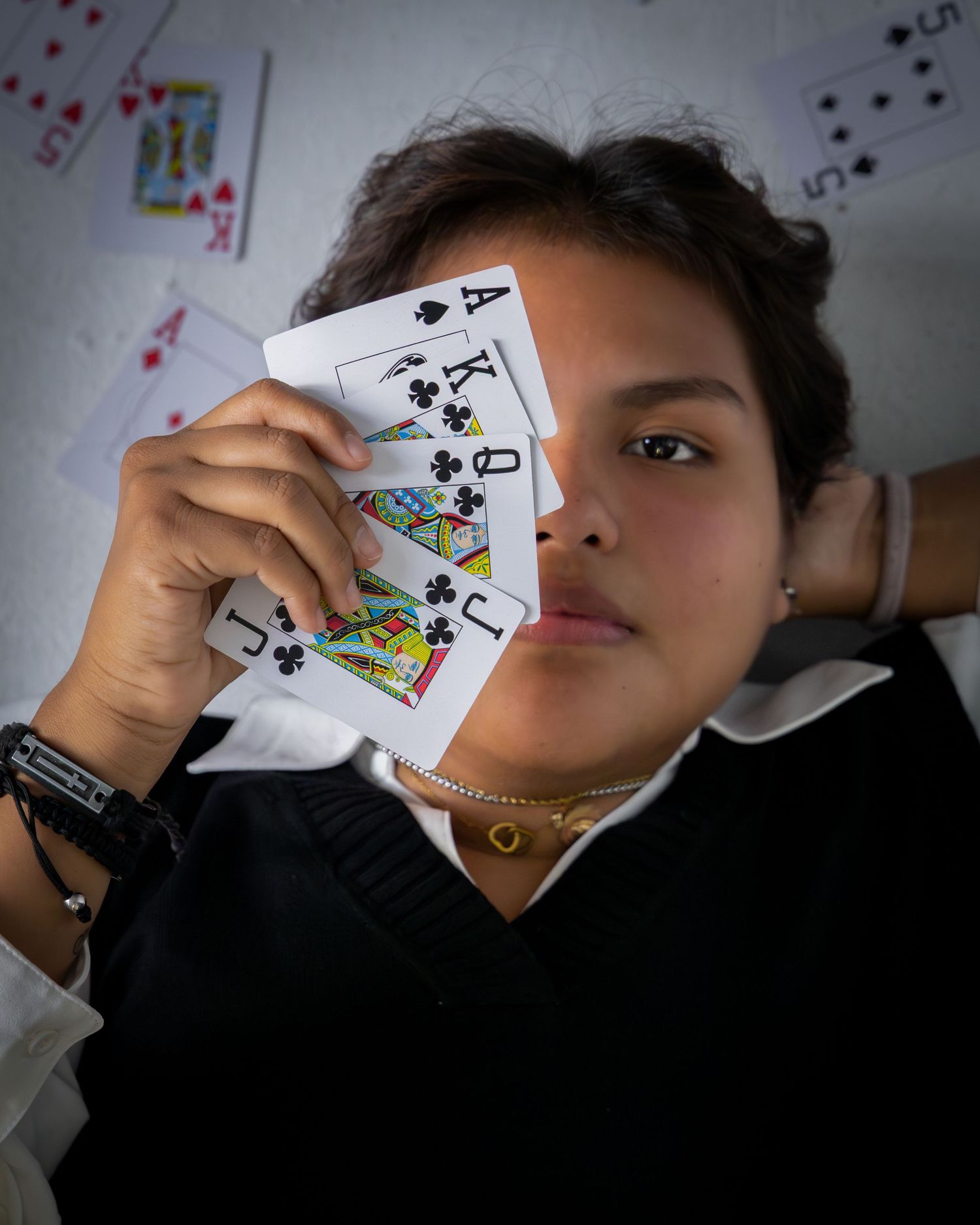 Someone holding playing cards in front of their face while wearing jewelry and a white shirt against a background with cards.