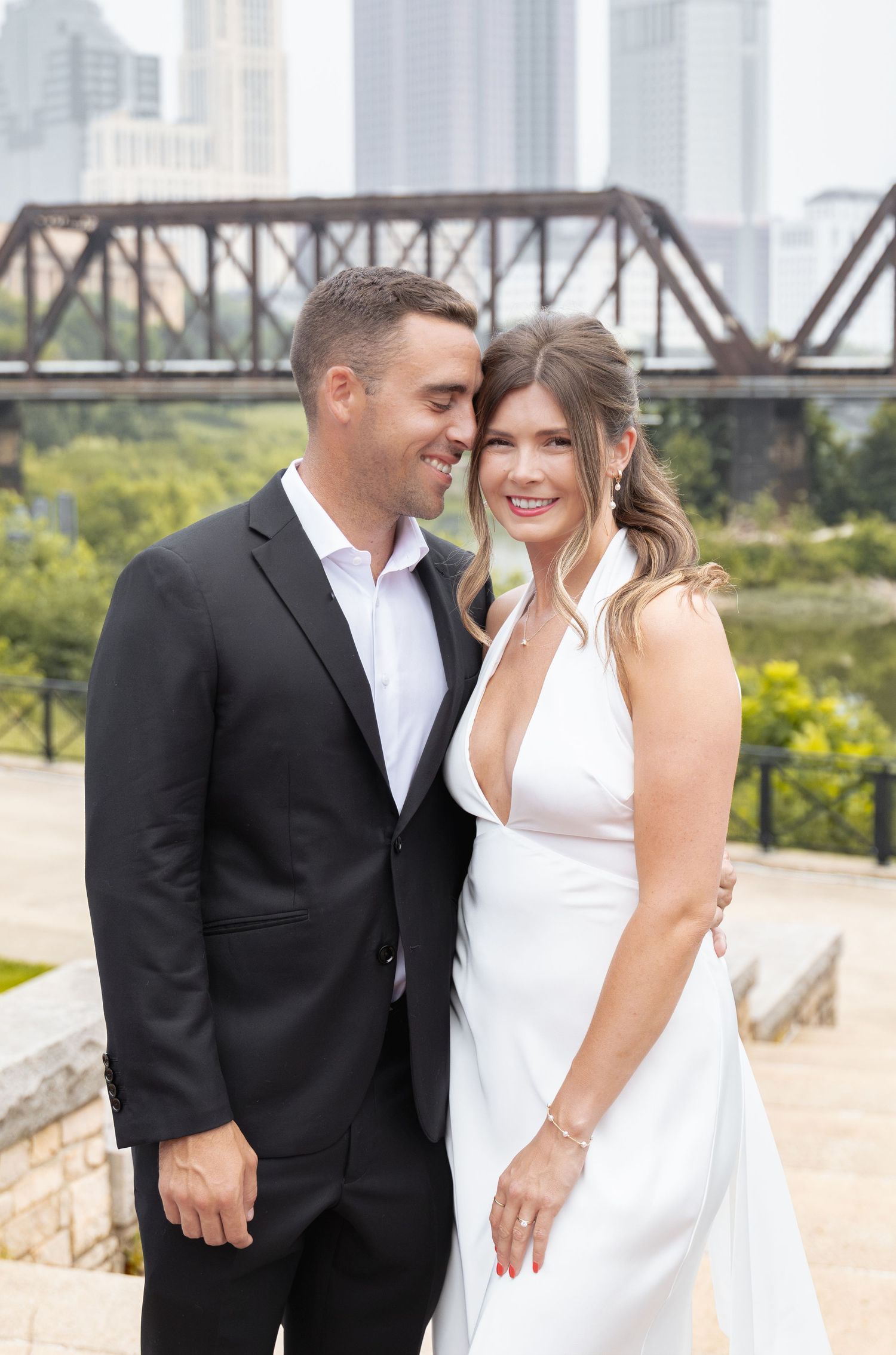 A couple in wedding attire smiles together outdoors with a steel bridge and city skyline in the background.