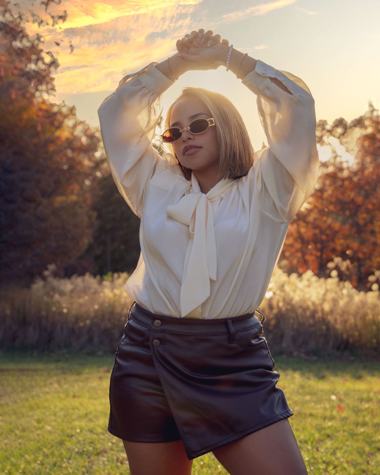 Woman in white blouse and leather shorts poses outdoors at golden hour sunset with arms raised.