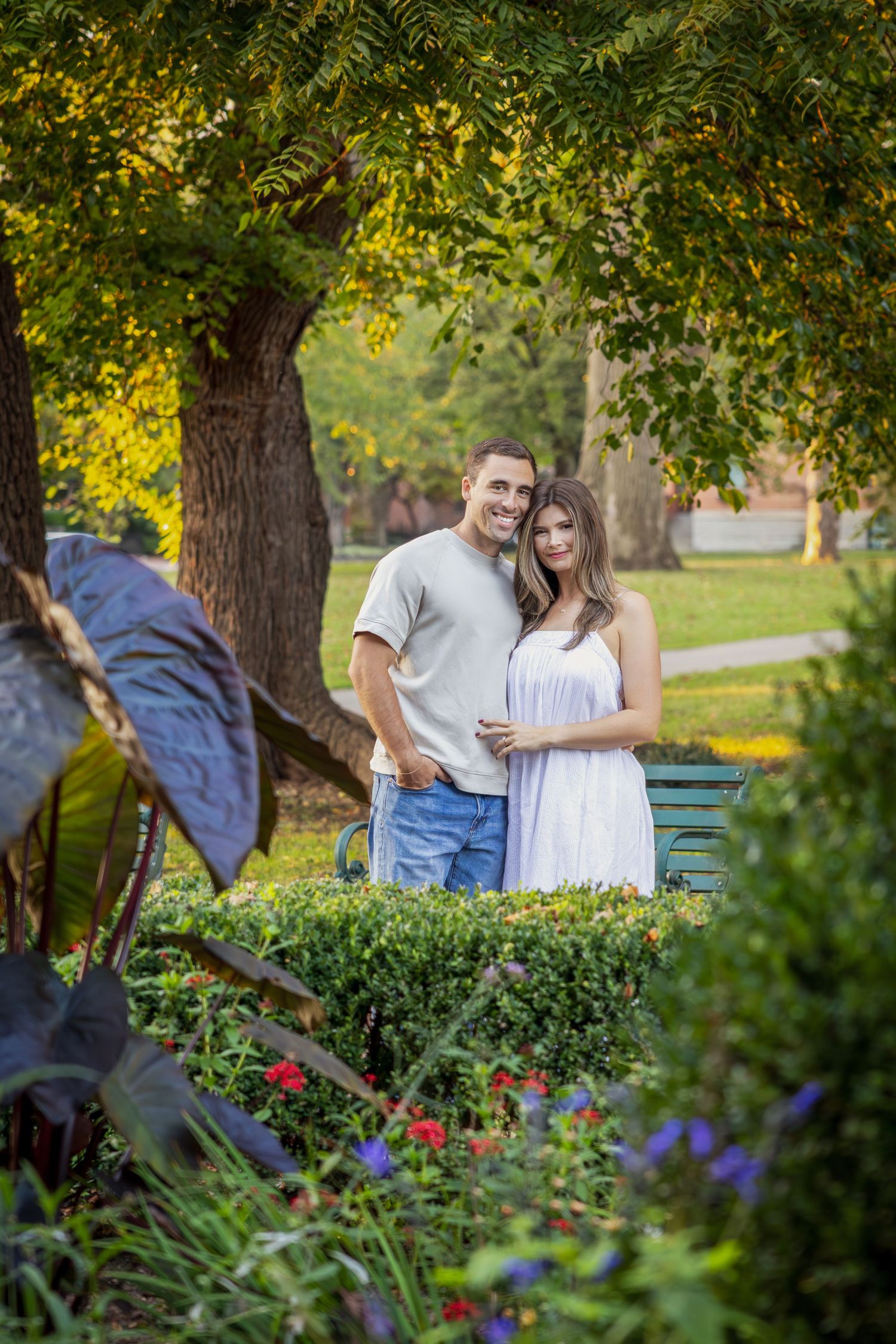 A smiling couple stands together in a lush green park garden, man in gray tee and jeans, woman in white dress.