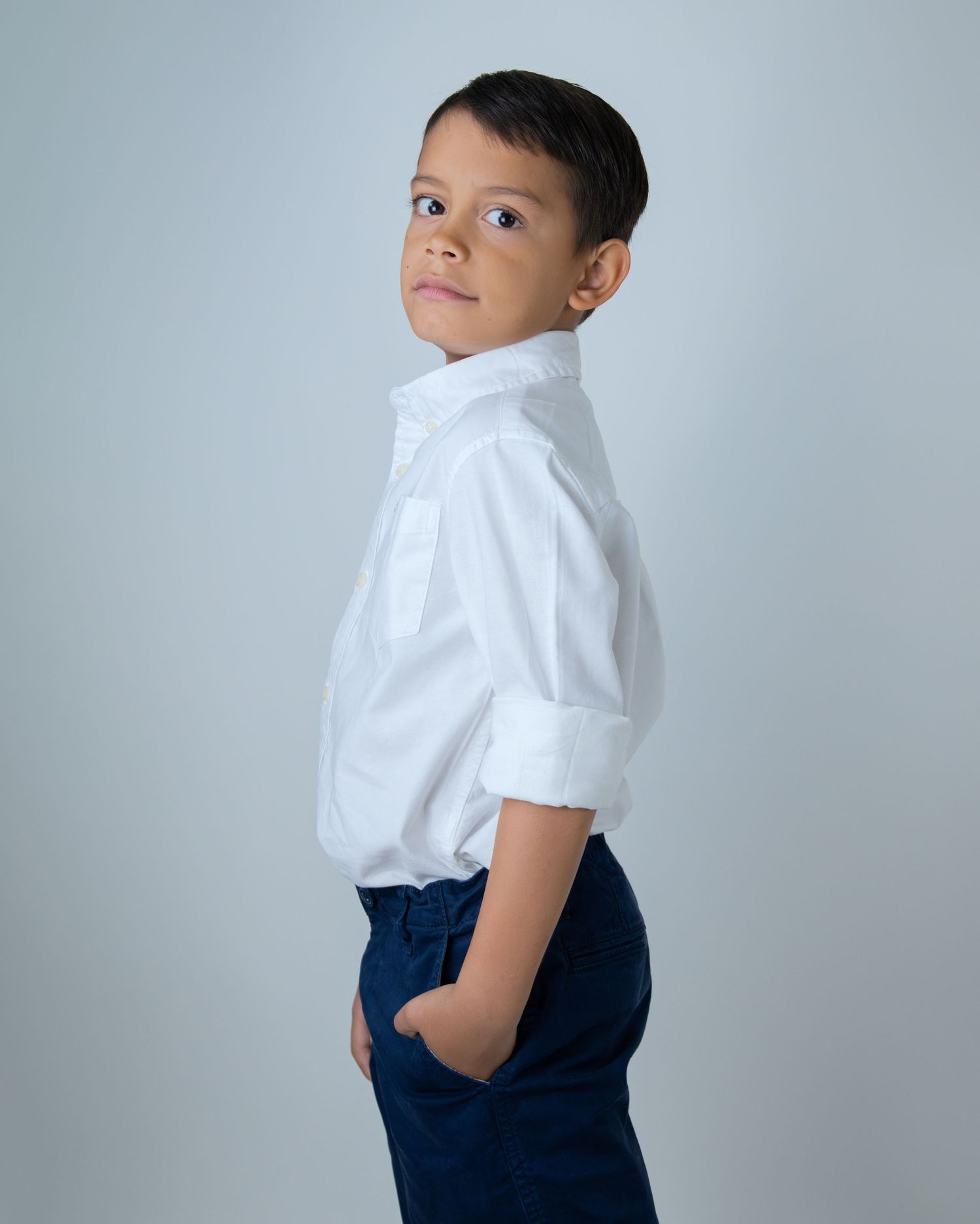 Young boy in white dress shirt and navy pants poses sideways, glancing over shoulder against gray background.