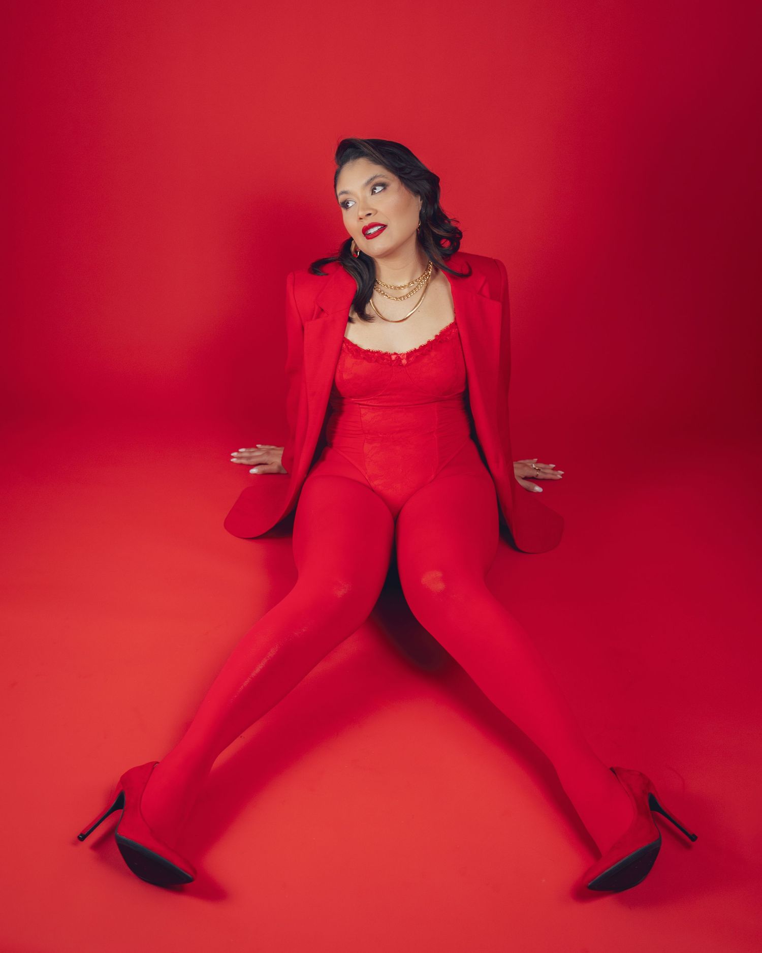 Woman in all-red outfit with blazer, bodysuit, and tights poses on red background wearing black heels.