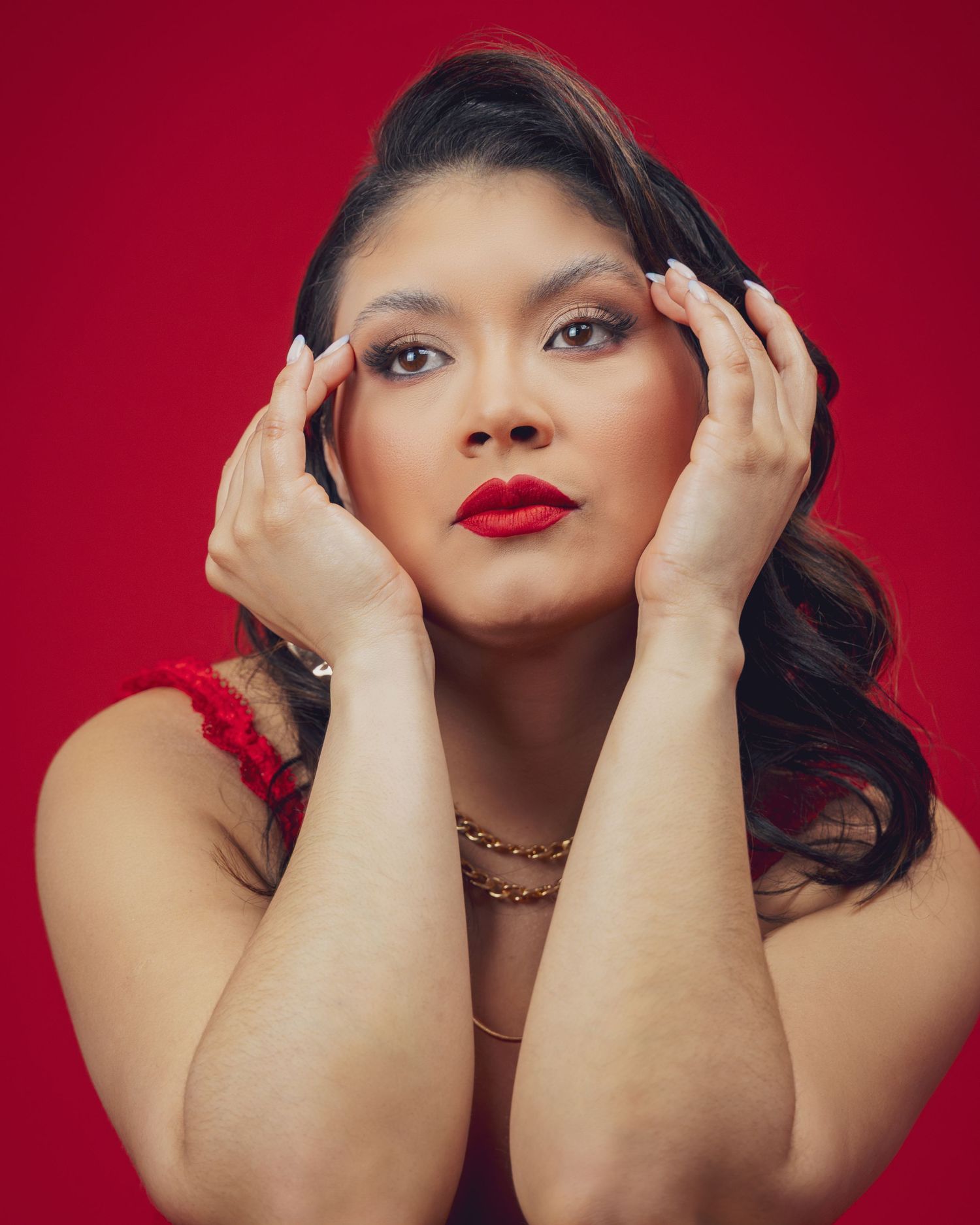 Woman with red lips and gold necklace poses against red background with hands framing her face.