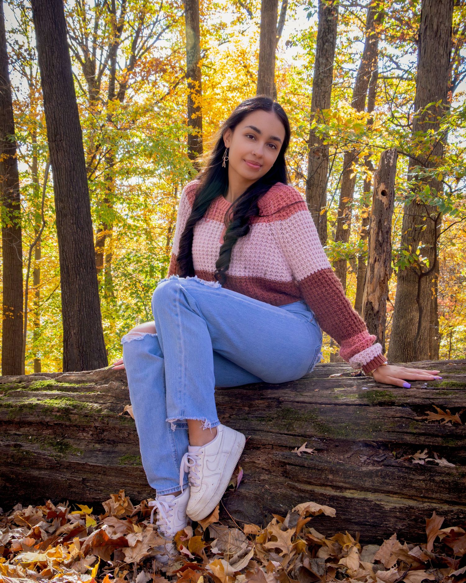 Young woman in pink sweater and jeans sitting on a log in an autumn forest with golden foliage.
