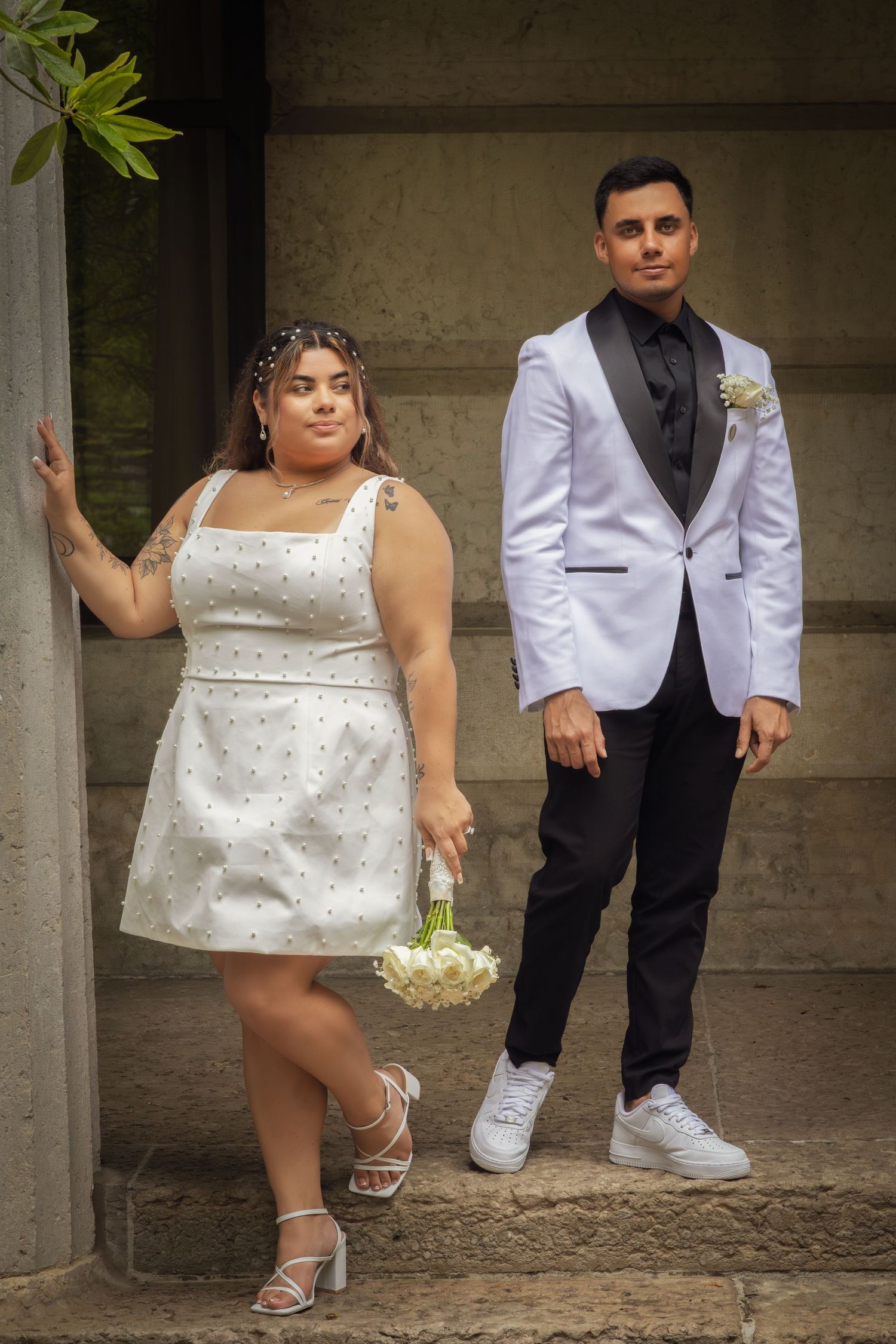 Bride in white mini dress holding bouquet poses with groom in white tuxedo jacket outdoors.