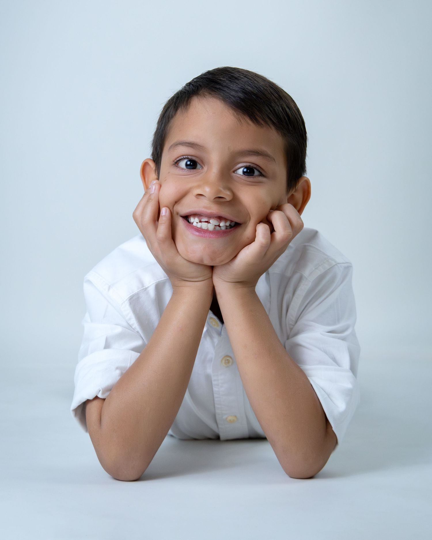 A smiling young boy in a white shirt rests his chin on his hands against a light gray background.