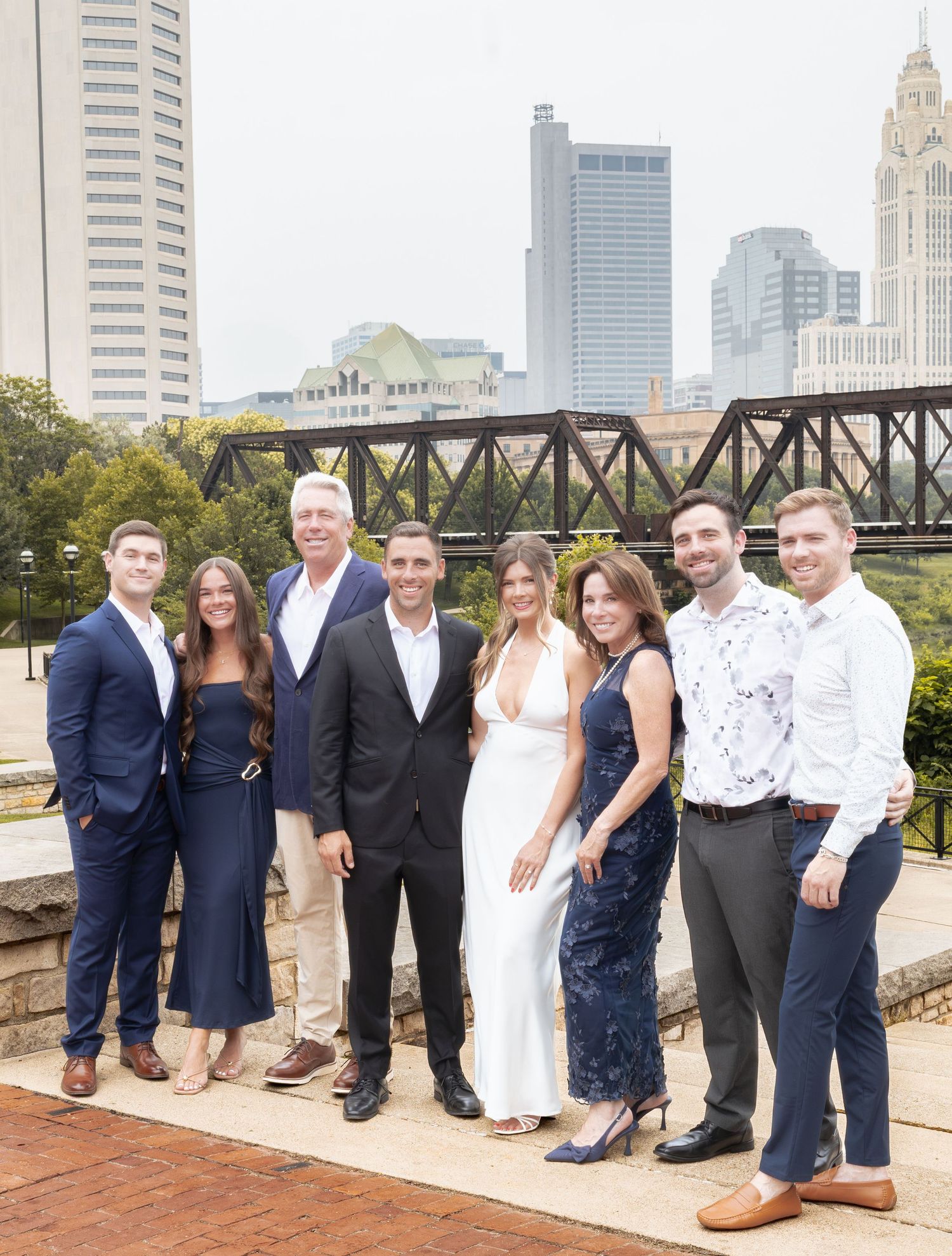 Group of seven adults posing outdoors with city skyline and bridge in background on a sunny day.