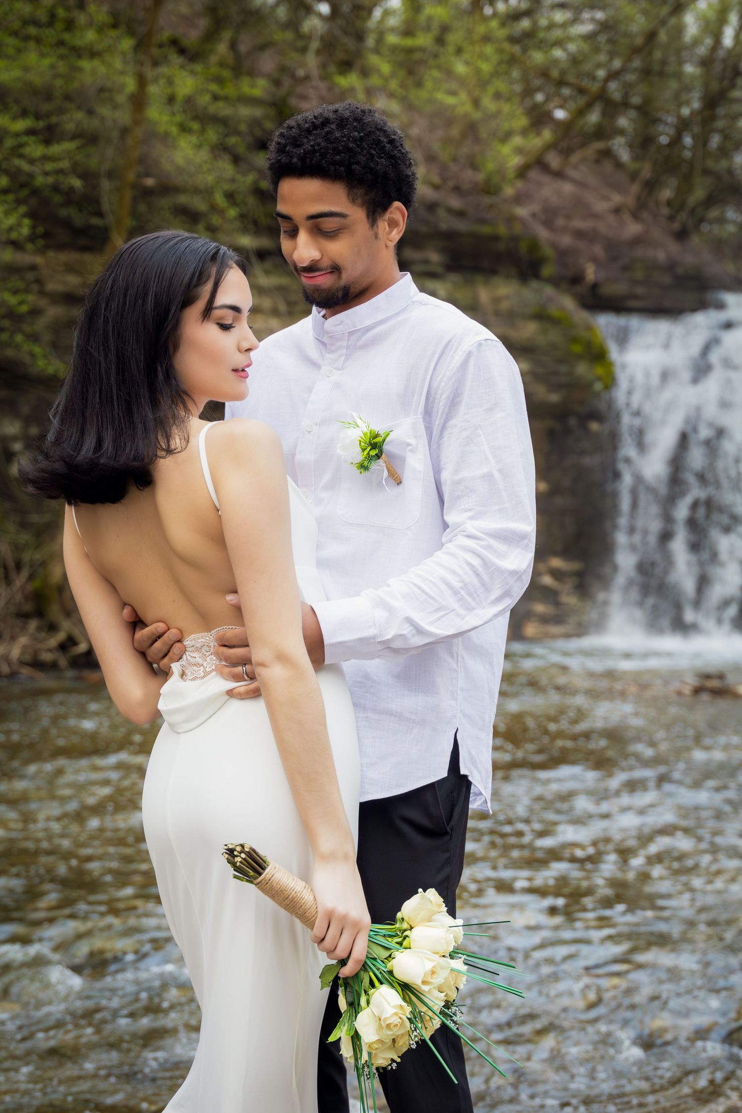 Interracial couple in wedding attire pose before a waterfall, bride in backless gown holding white roses bouquet.