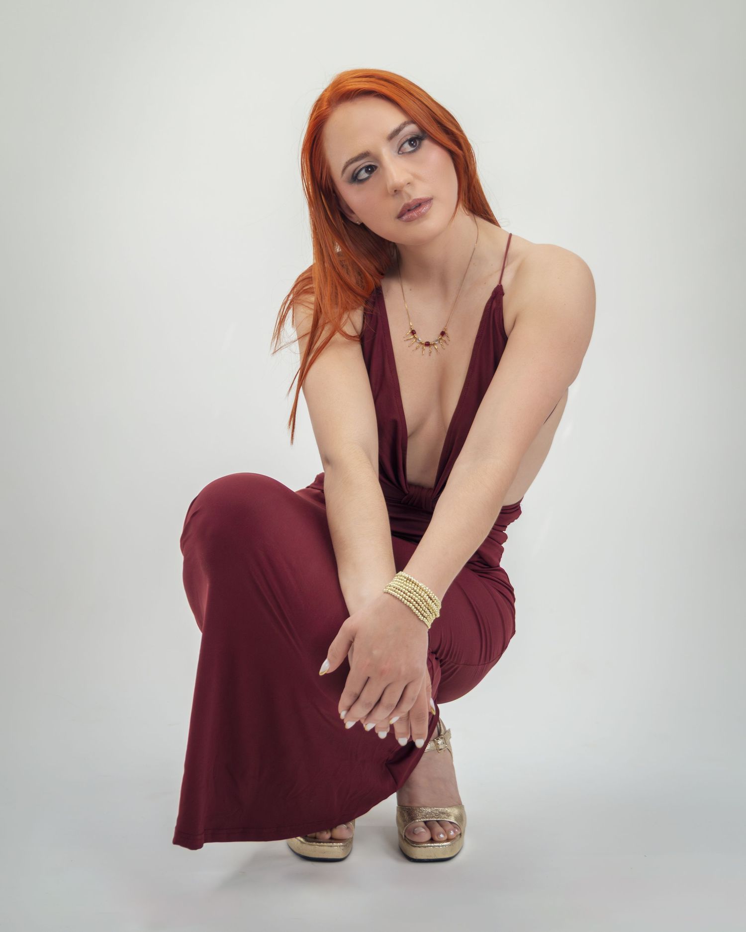 A young woman with long red hair poses in a deep V-neck burgundy jumpsuit and gold heels, crouching gracefully.