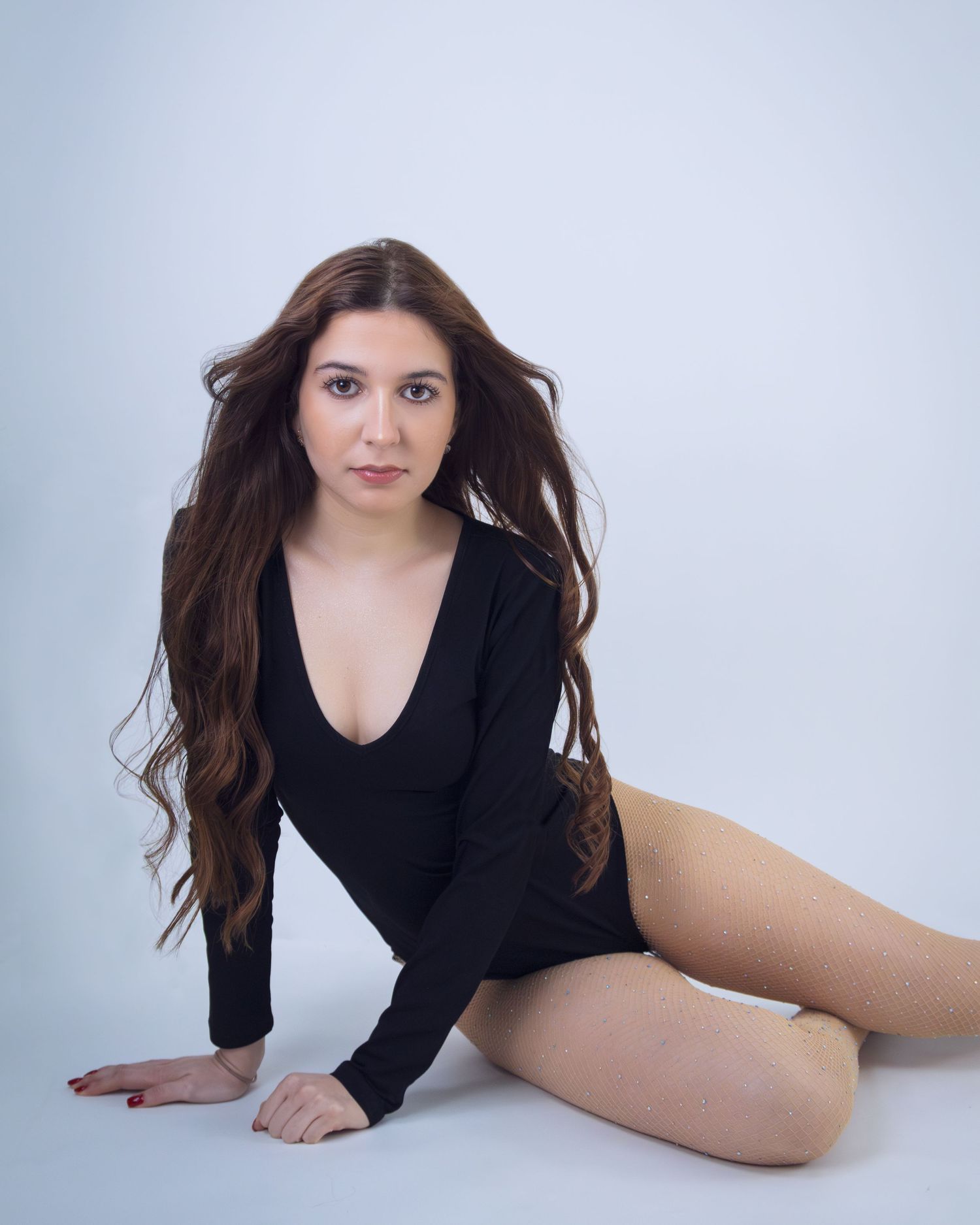 Young woman with long wavy brown hair poses in a black bodysuit against a white studio background.