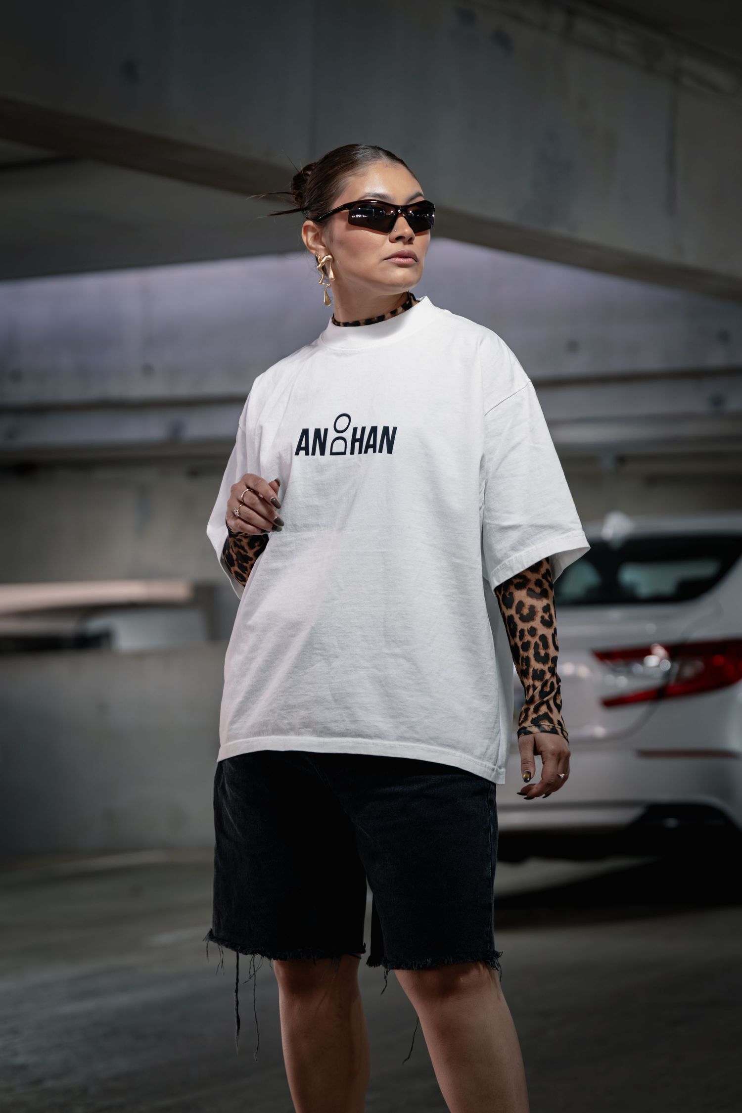 Person wearing white t-shirt with Anshan text and black shorts walking in parking garage with tattoo sleeve visible.