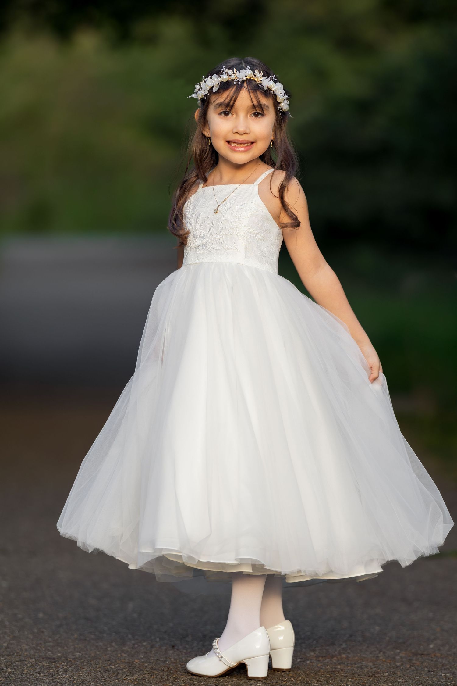 Young girl in white tulle dress and floral crown poses gracefully outdoors for what appears to be a special occasion.