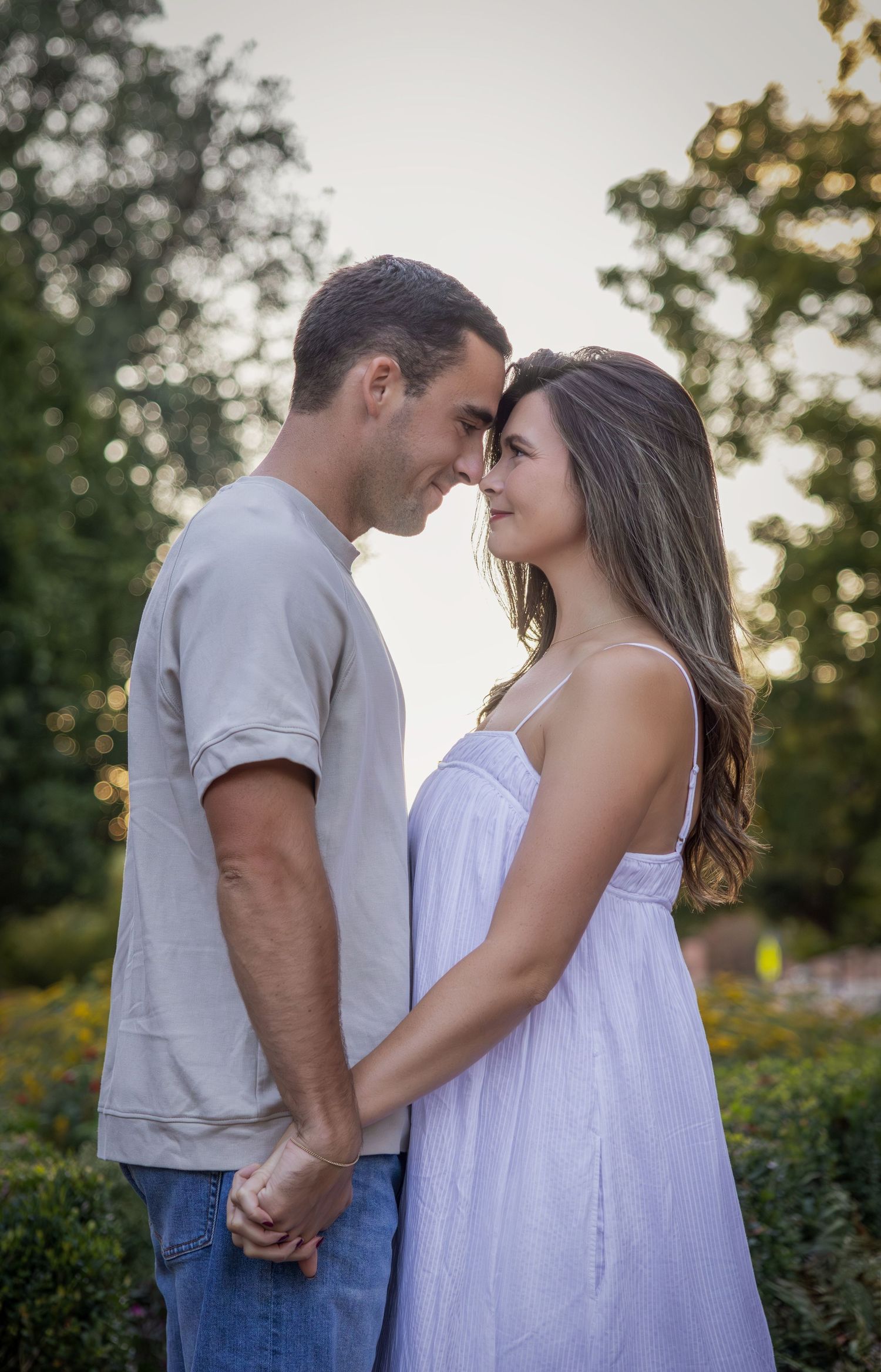 A couple stands forehead-to-forehead holding hands in a lush garden at dusk, sharing a tender romantic moment.