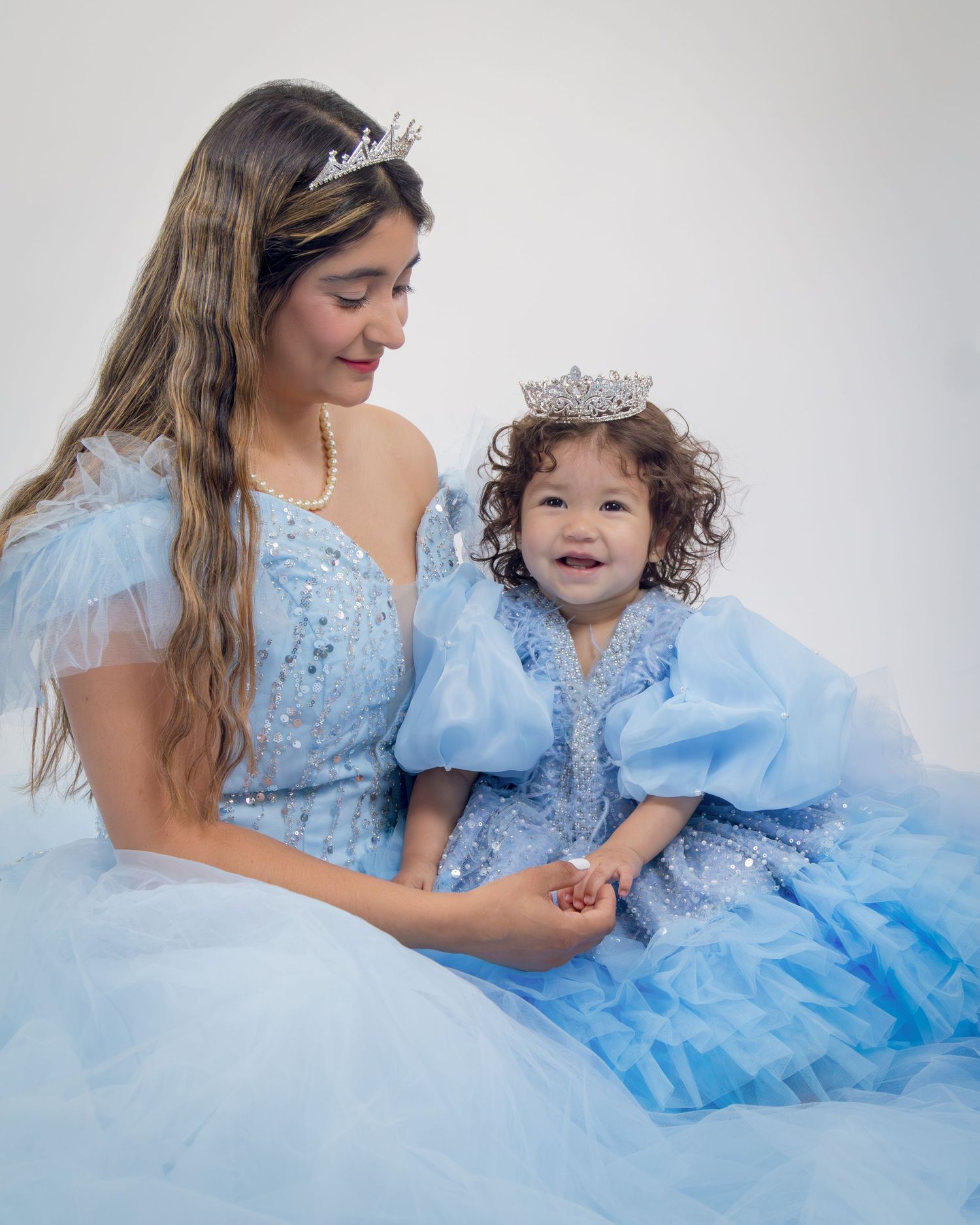 Mother and toddler in matching sparkly blue princess gowns and crystal tiaras pose together in a studio photo.