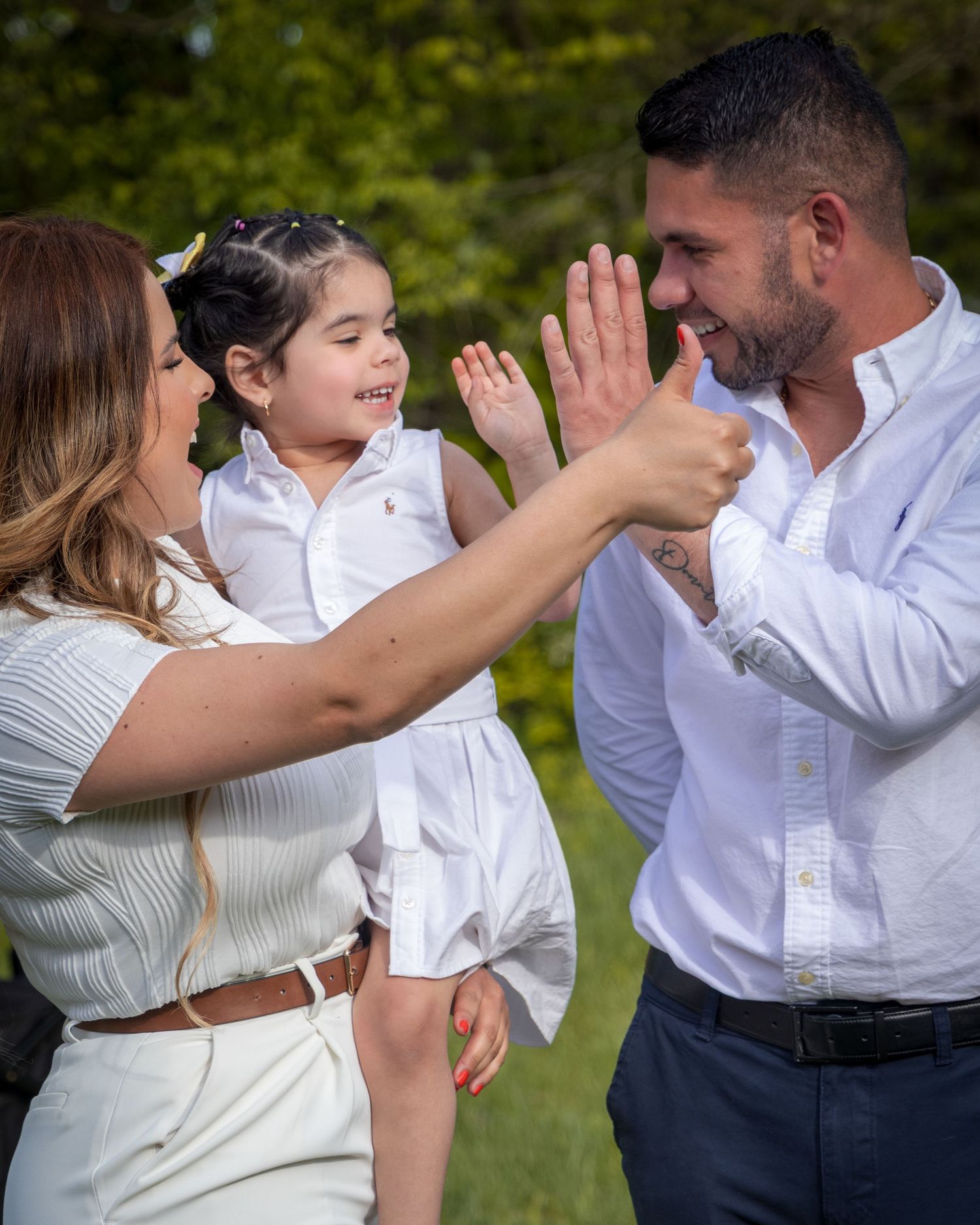 A joyful family in white outfits shares a high-five outdoors, with parents smiling at their young daughter.