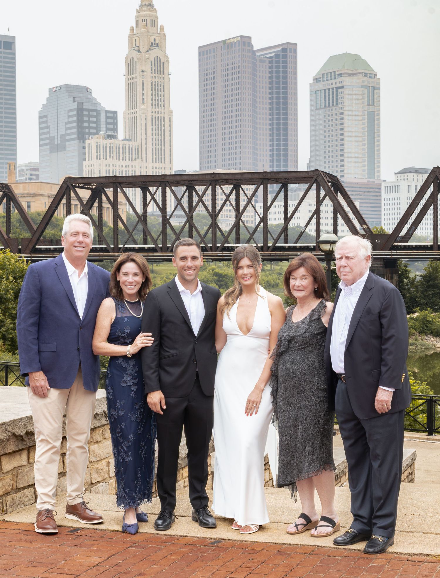 Group of six people posing together outdoors with a city skyline and iron bridge in the background.