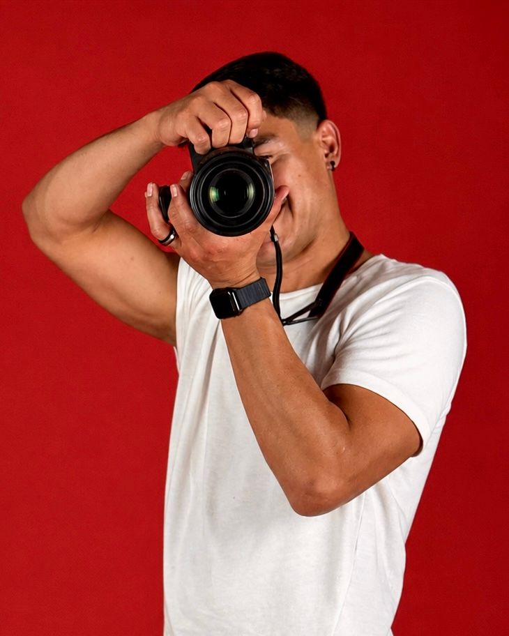 Man in white t-shirt photographing with DSLR camera against red background.