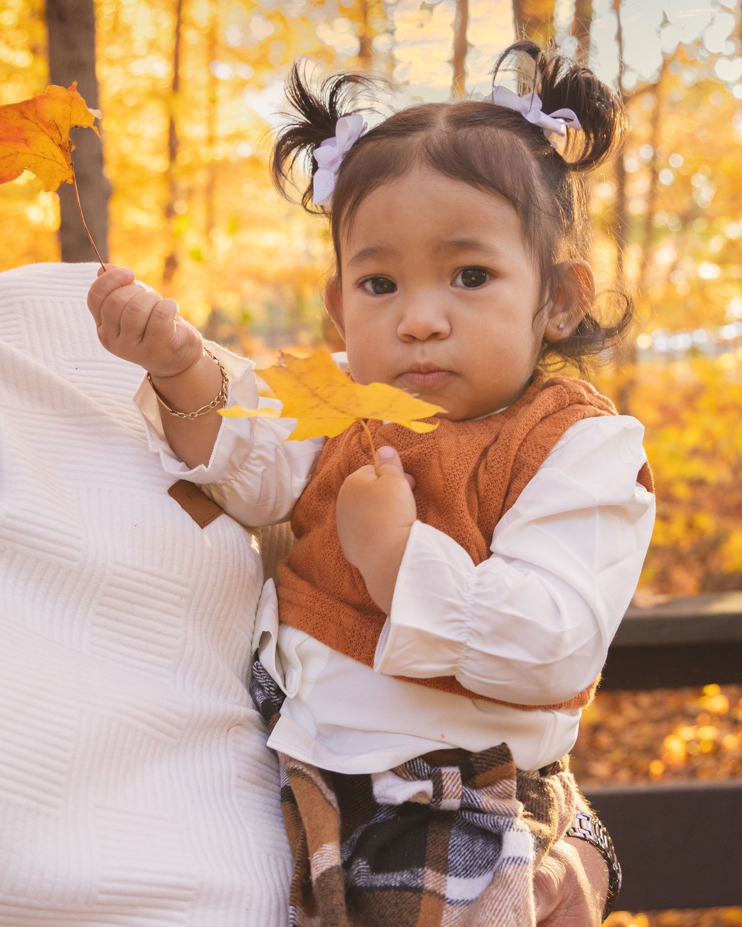 Toddler girl in autumn vest holds yellow maple leaf outdoors among golden fall foliage.