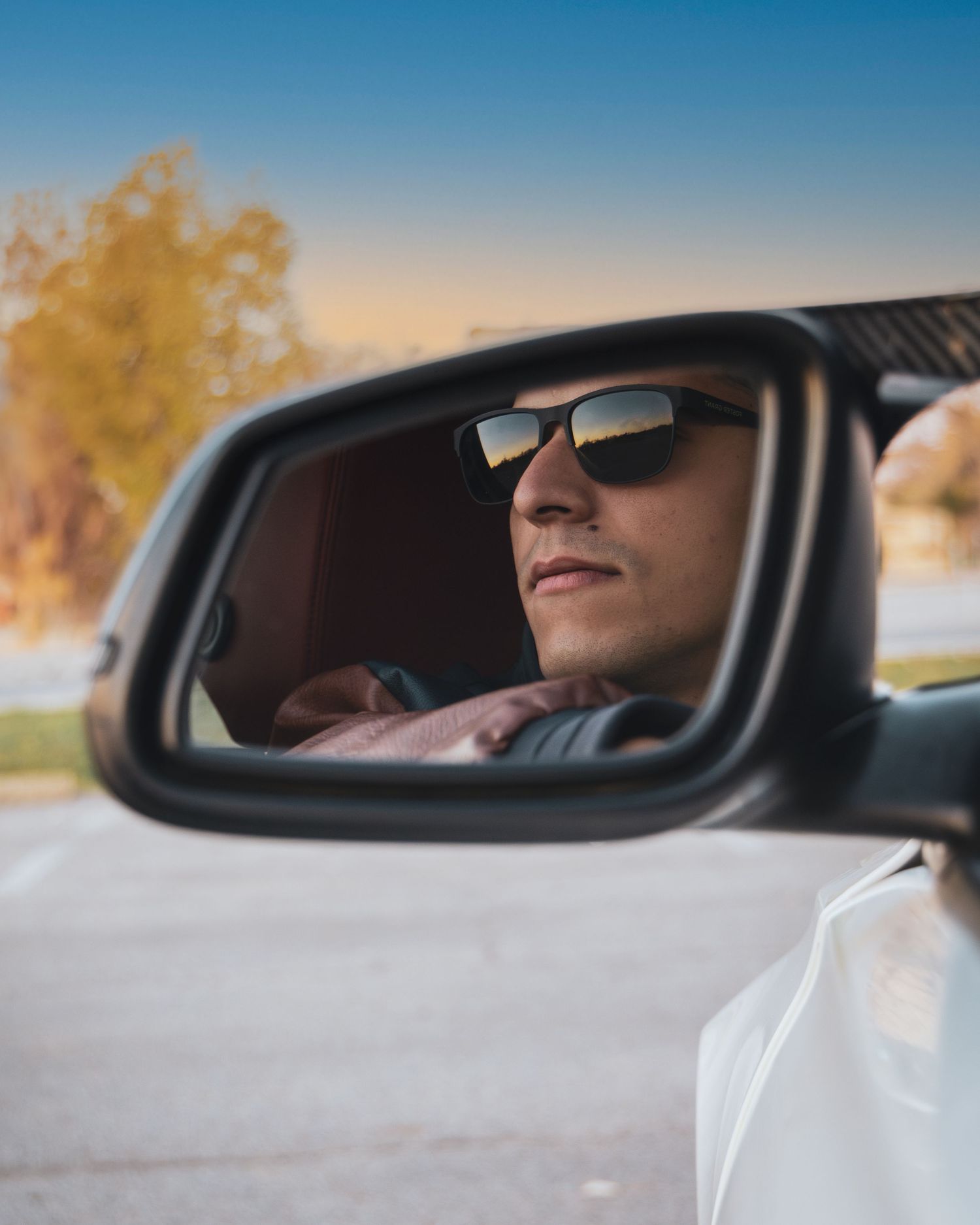 A man wearing sunglasses reflected in a car side mirror on a sunny day with trees in the background.