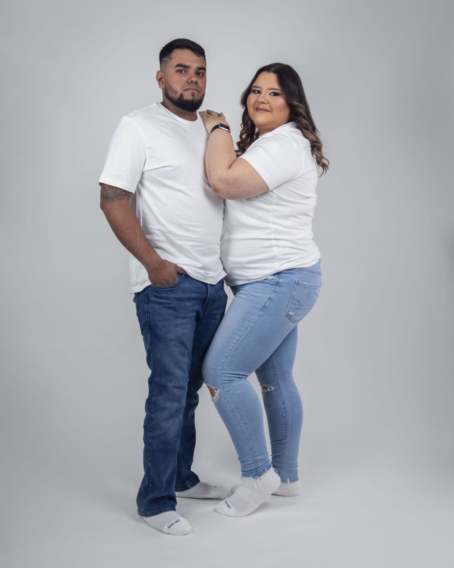 A couple in matching white t-shirts and jeans posing together against a light gray studio background.