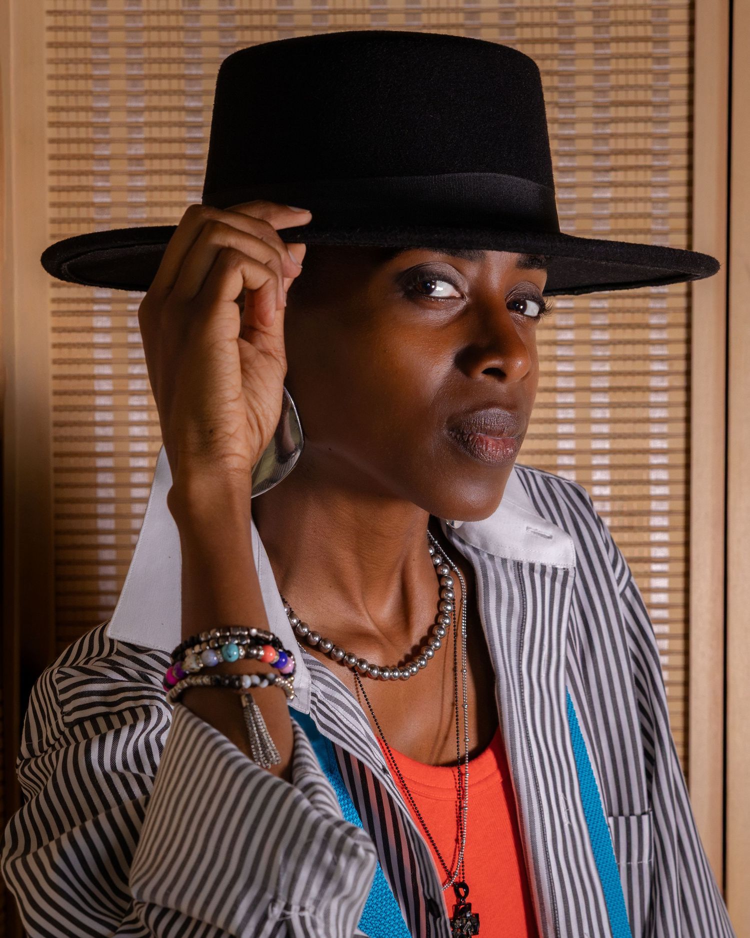 Woman in a black wide-brim hat tips the brim, wearing a striped jacket, orange top, beaded necklaces, bracelets and large hoop earrings.