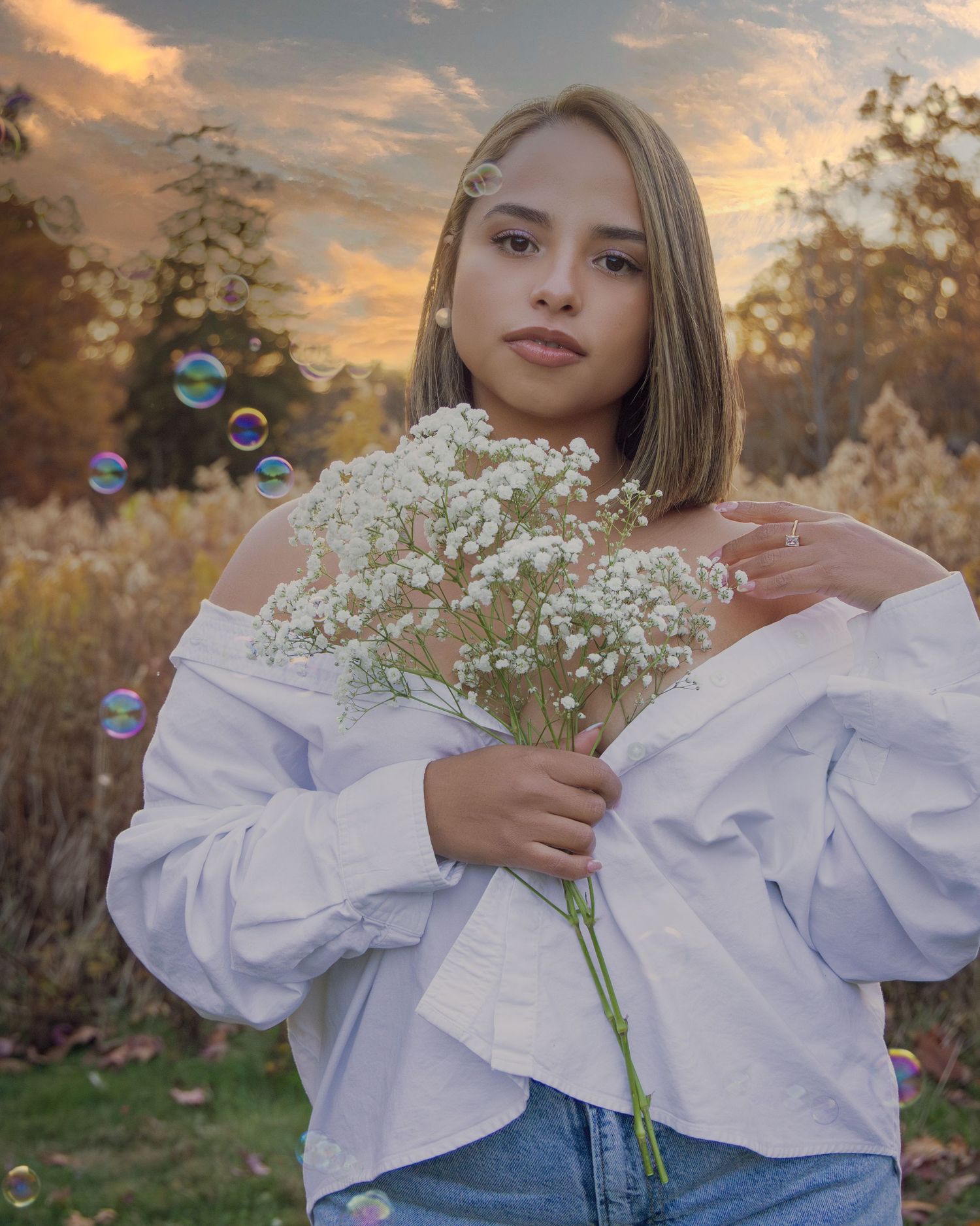 Young woman in white off-shoulder top holds white baby's breath flowers outdoors at sunset with colorful bubbles floating around her.